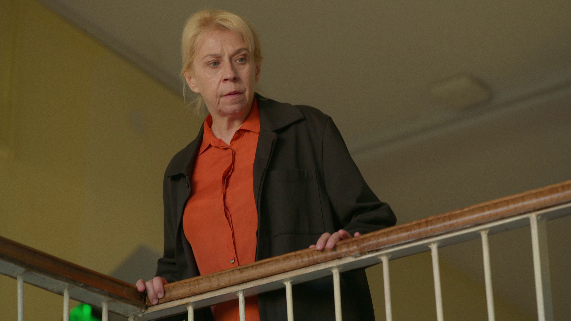 A woman with blonde hair, wearing an orange shirt and a black jacket, looks down with a concerned expression while standing behind a wooden railing.