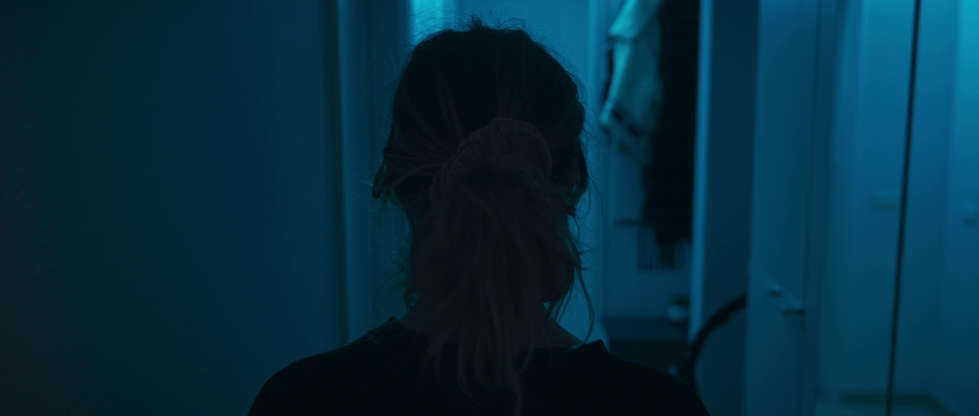 Back view of a woman with curly hair tied in a ponytail, walking through a dimly lit corridor with blue lighting, surrounded by lockers.