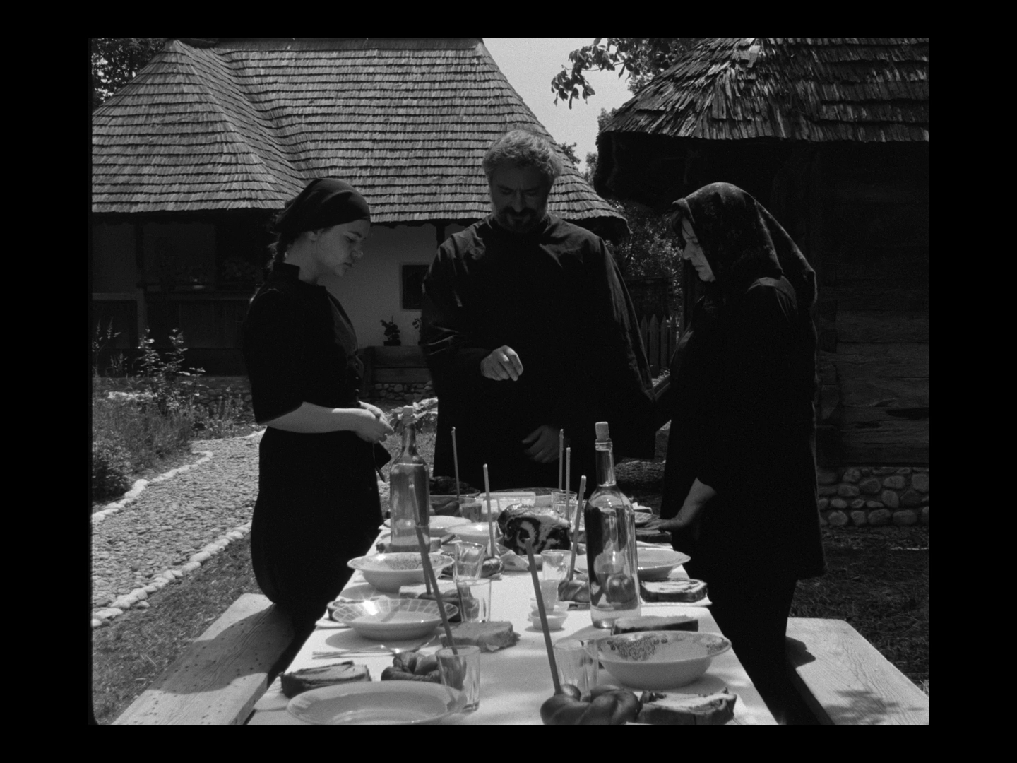 Three people standing around a table outdoors with food and drinks, in front of traditional wooden houses with shingled roofs.