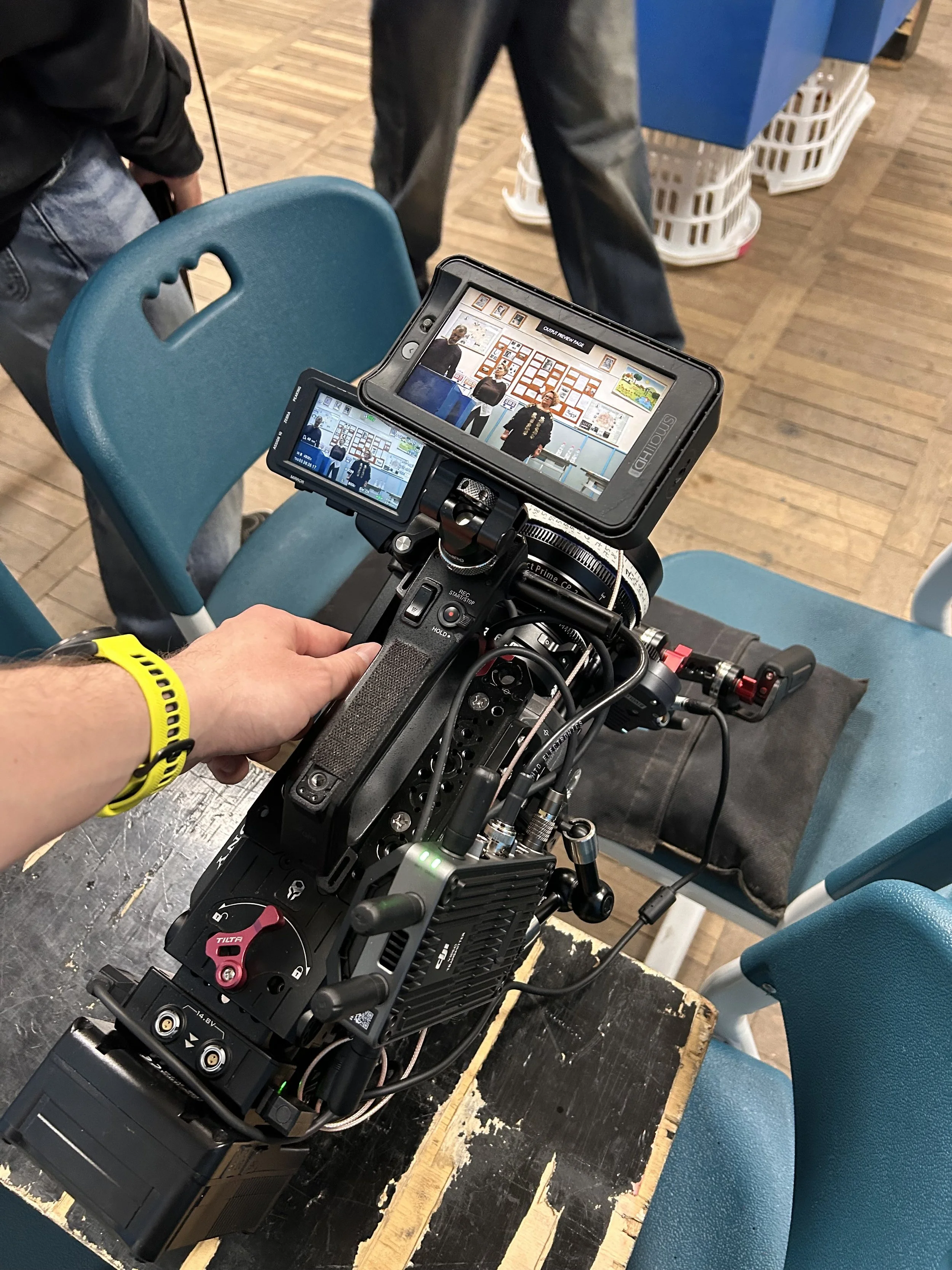 Close-up of a professional video camera on a tripod capturing a scene of two people standing in front of a wall decorated with framed pictures.