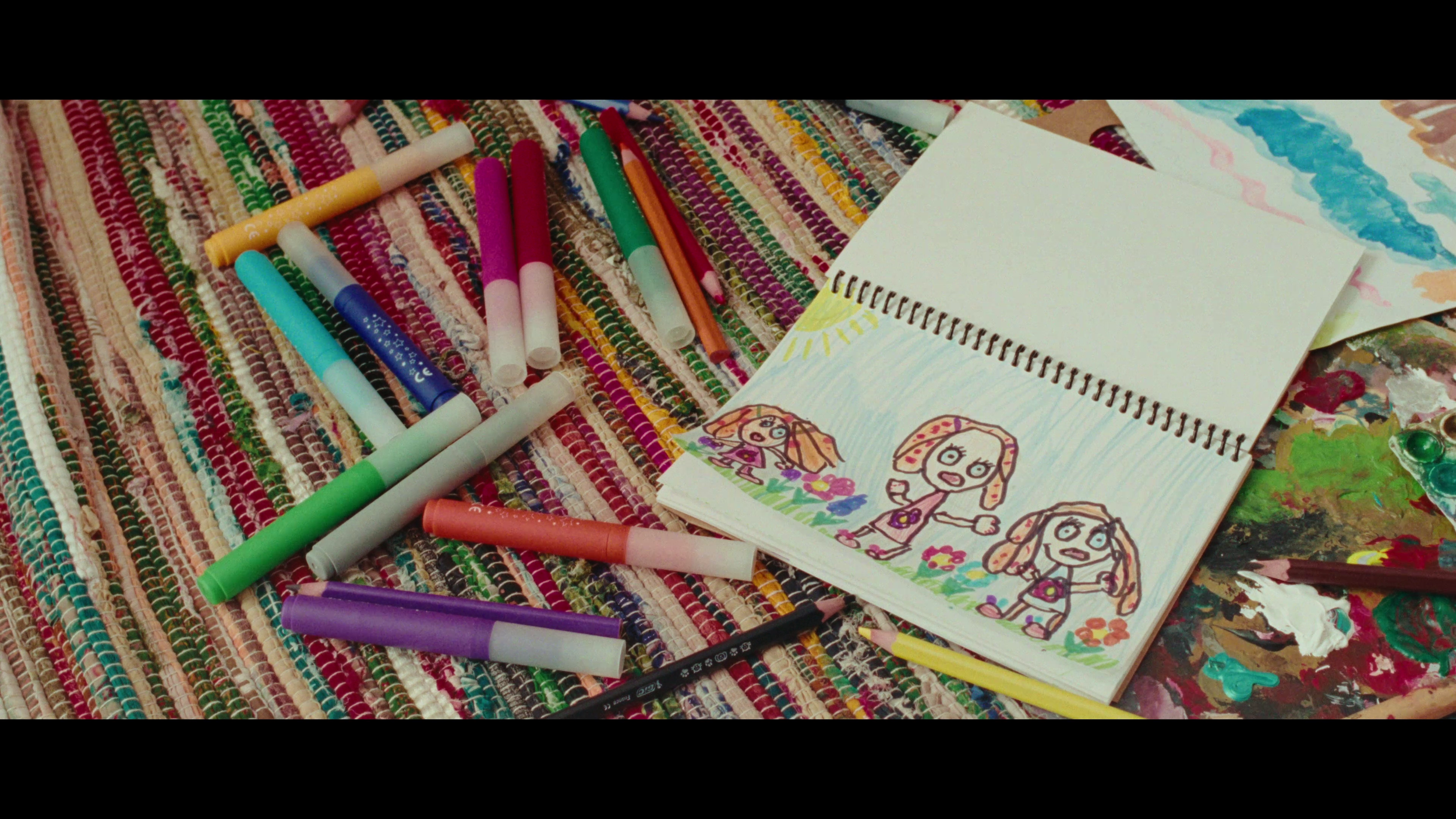 A colorful woven rug with markers, a drawing pad, and child's crayon and marker drawings, featuring three cartoon girl characters in a grassy field with a bright sun.