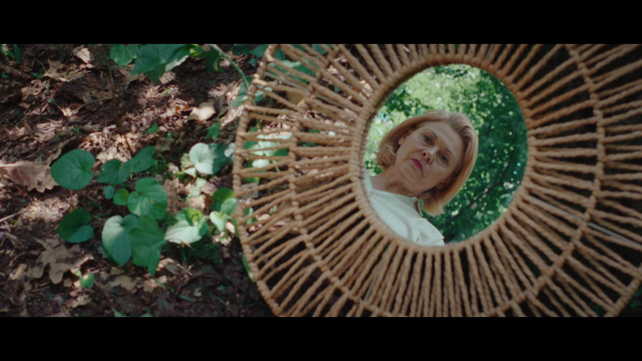 A woman with blonde hair looking down at a small mirror placed on the ground in a forest with green foliage.