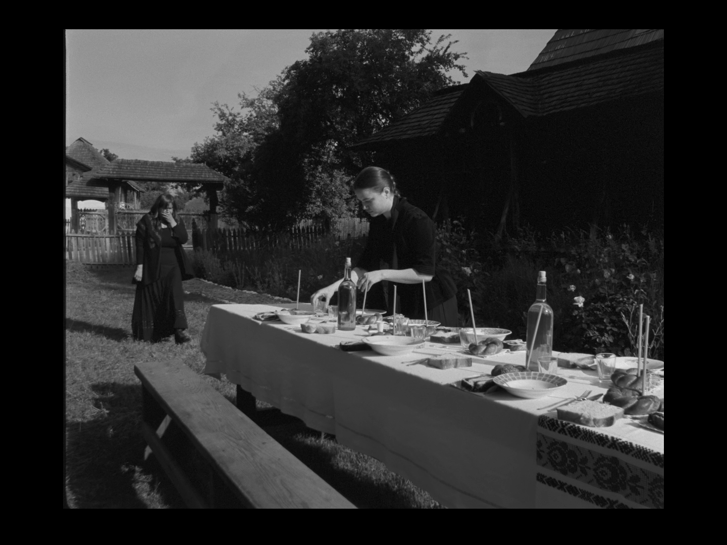 A woman arranging food on a table outdoors in daytime, with another woman walking towards her in the background. The table has various dishes and bottles, set in a rural setting with a house, trees, and a wooden fence.