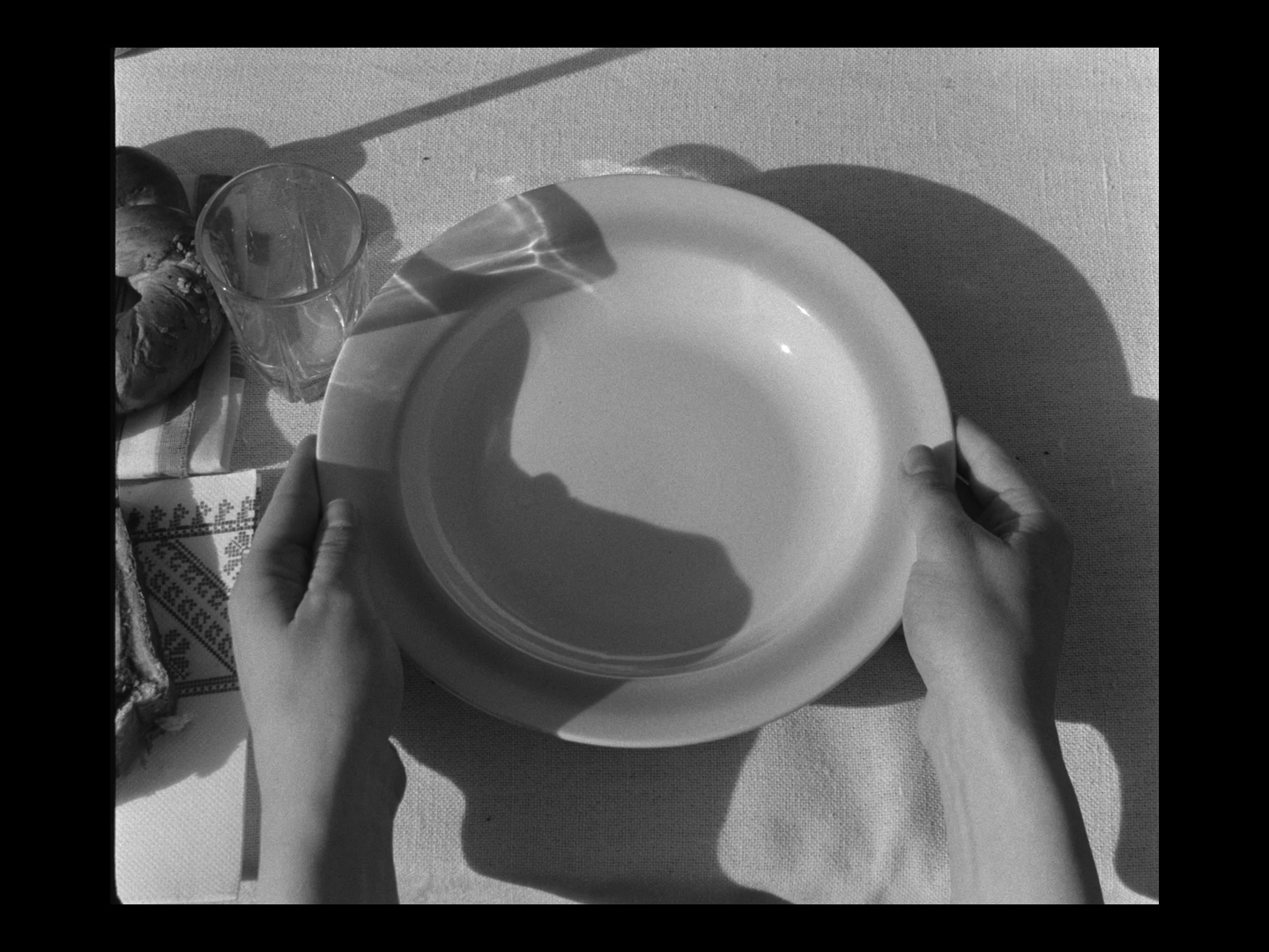 Black and white photo of two hands holding a white plate above a table with a glass of water and some baked goods.