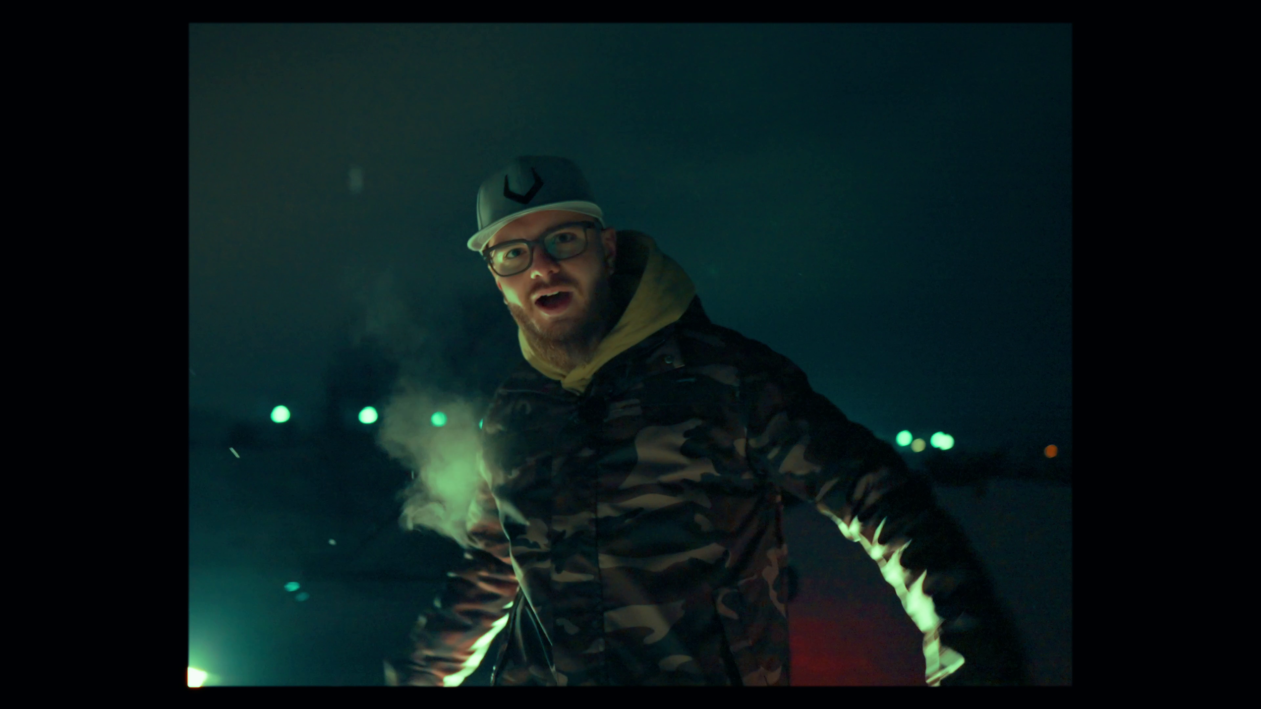 A man with glasses, a beard, and a surprised expression, wearing a gray cap with a logo, a camouflage hoodie, standing in a dark outdoor setting with green and orange lights in the background.