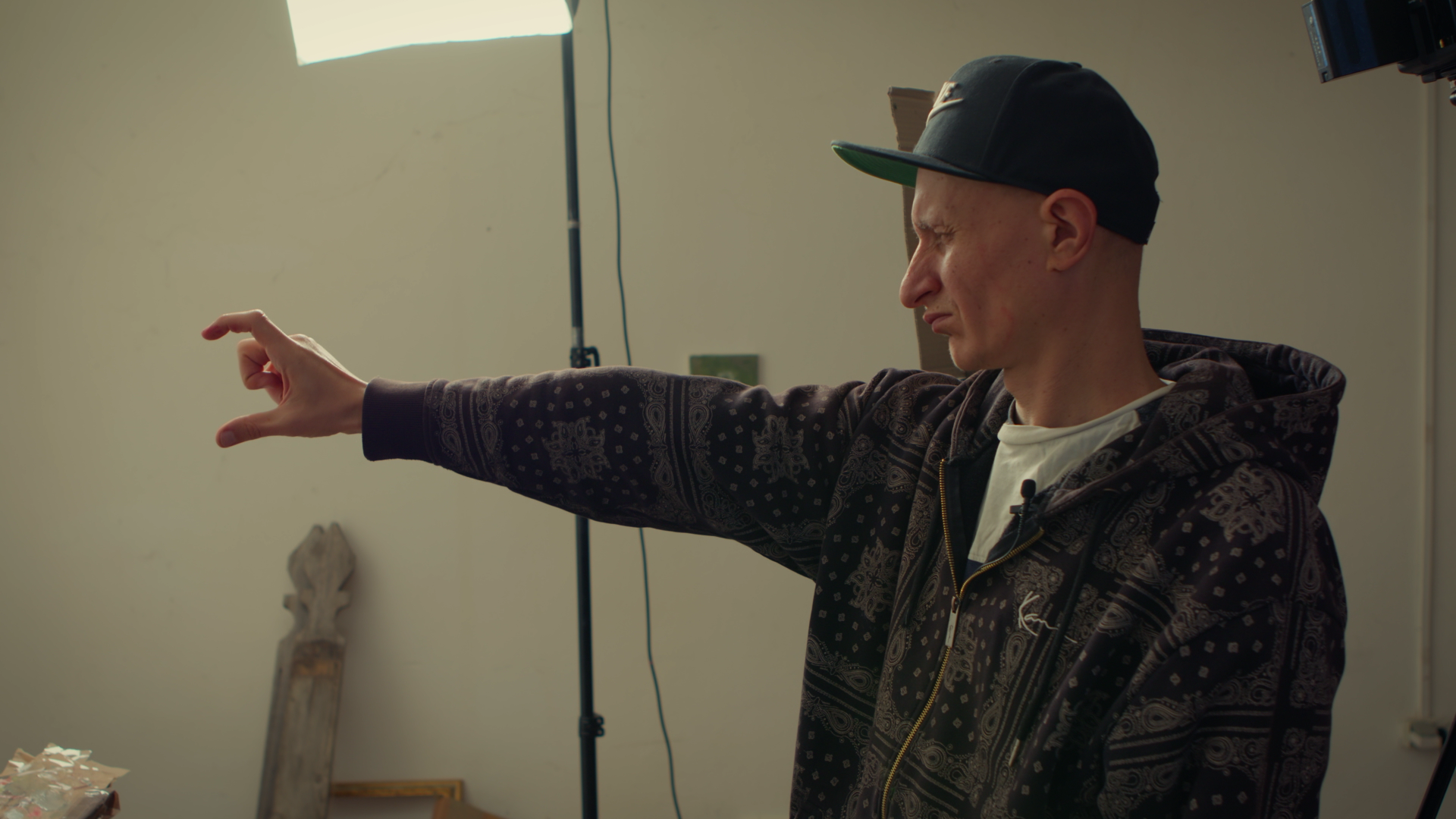 A man with a black cap, patterned hoodie, and white T-shirt is extending his right arm and making a gesture with his hand. He is looking intensely to the left, in an indoor setting with a light-colored wall, a bright light, and a camera or lighting equipment in the background.
