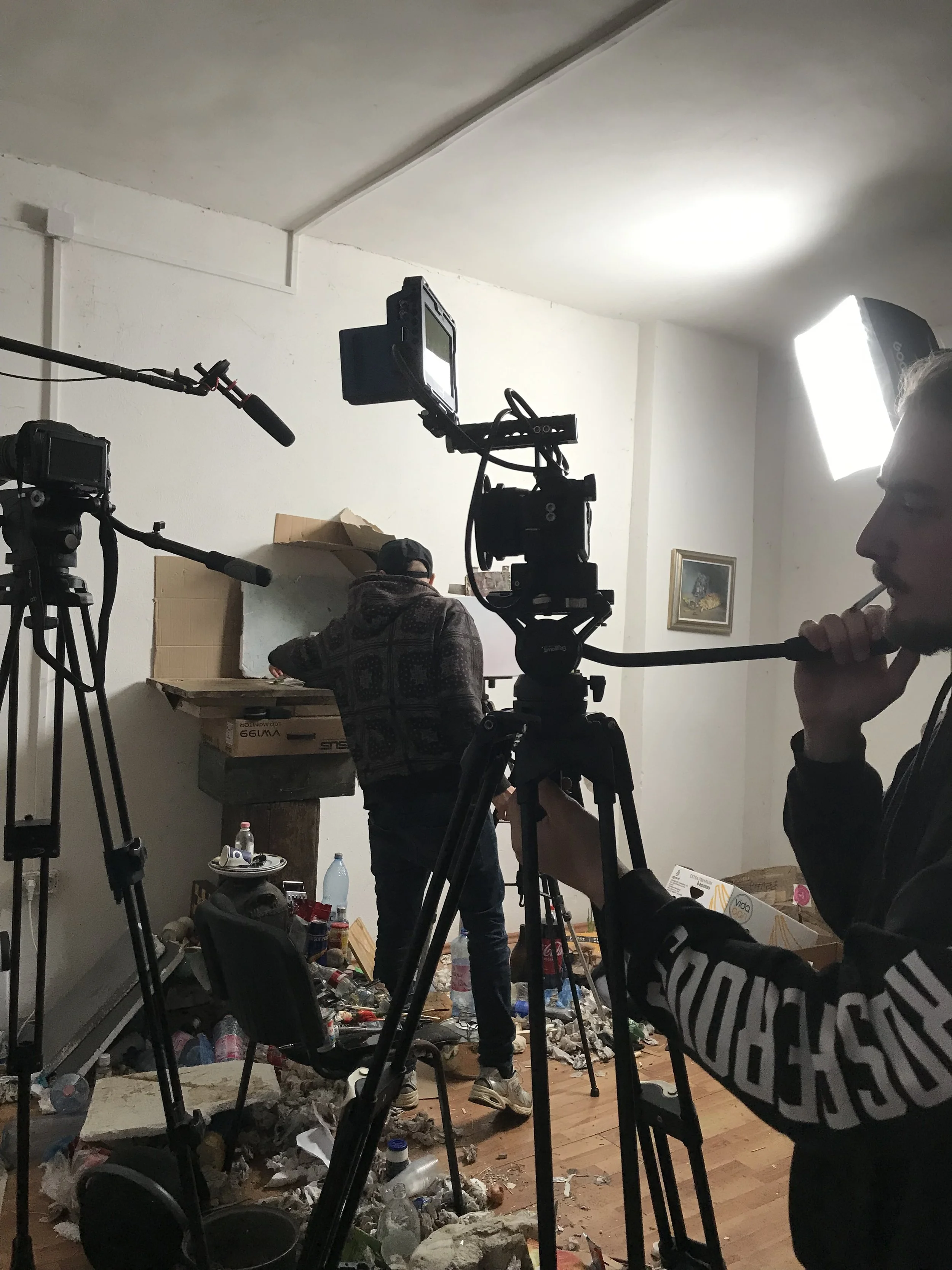 Filmmakers filming a scene in a cluttered room with trash, pizza boxes, and bottles scattered on the floor. One person is operating a camera, another is holding a boom microphone, and a third person is in the background organizing or adjusting items on a table.