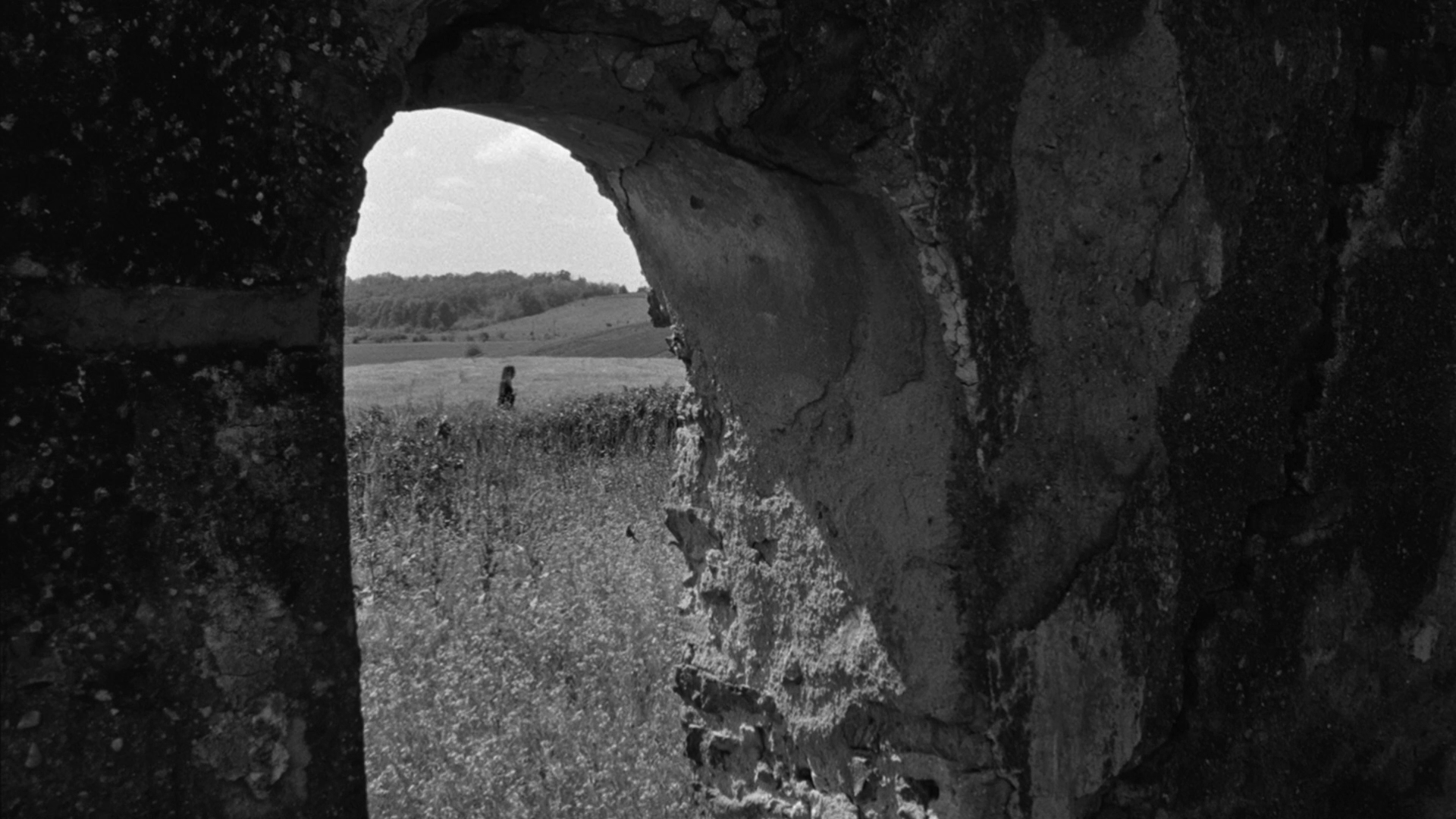 View through a stone window in a structure, looking out at a grassy field with a person walking and rolling hills in the distance.