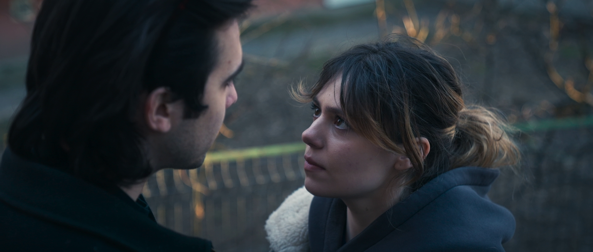 A man and woman face each other closely outdoors, with the man on the left, and the woman on the right, both with intense expressions.