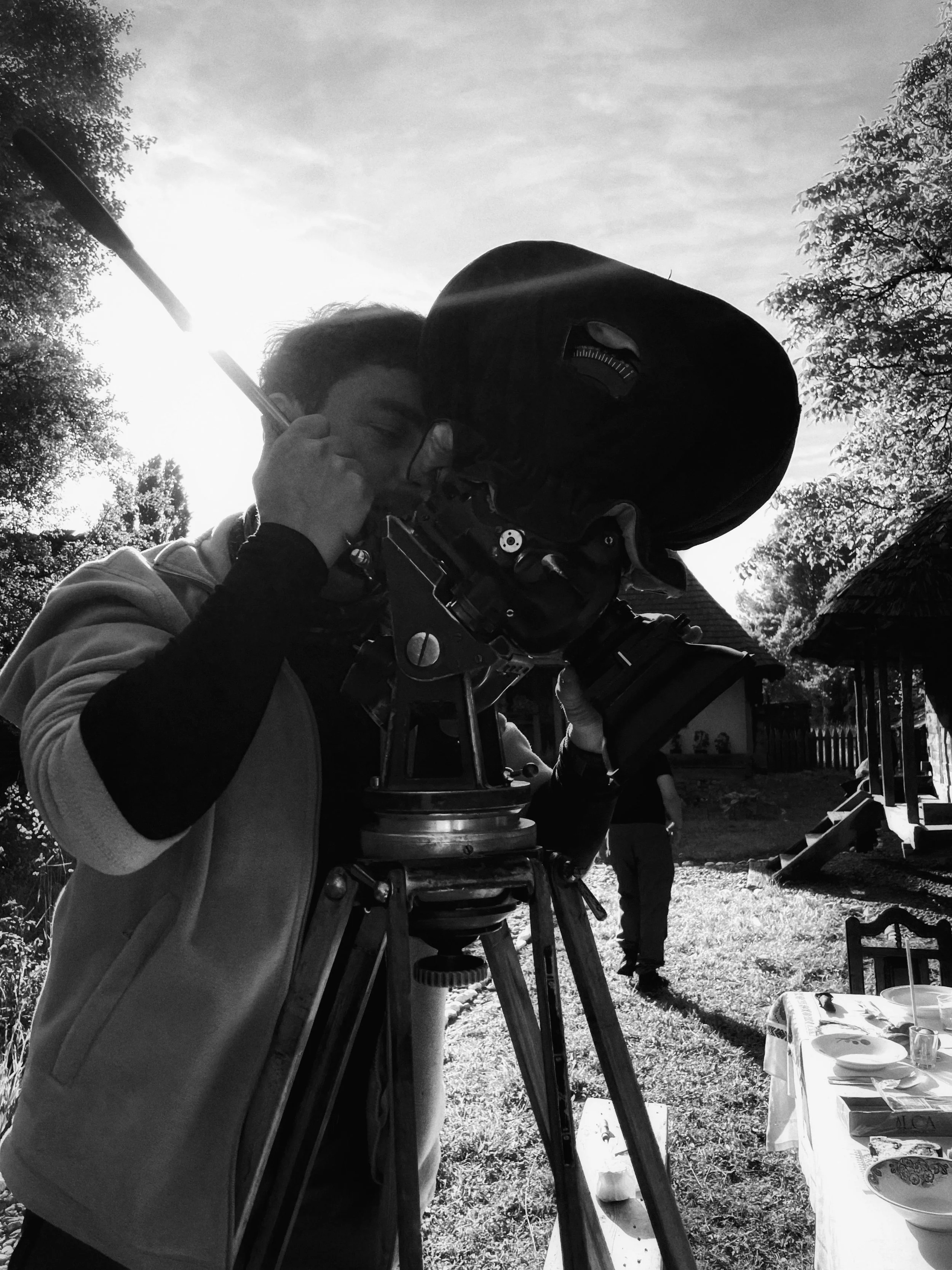 Person looking through a large telescope outdoor in sunlight with trees and a small building in the background.
