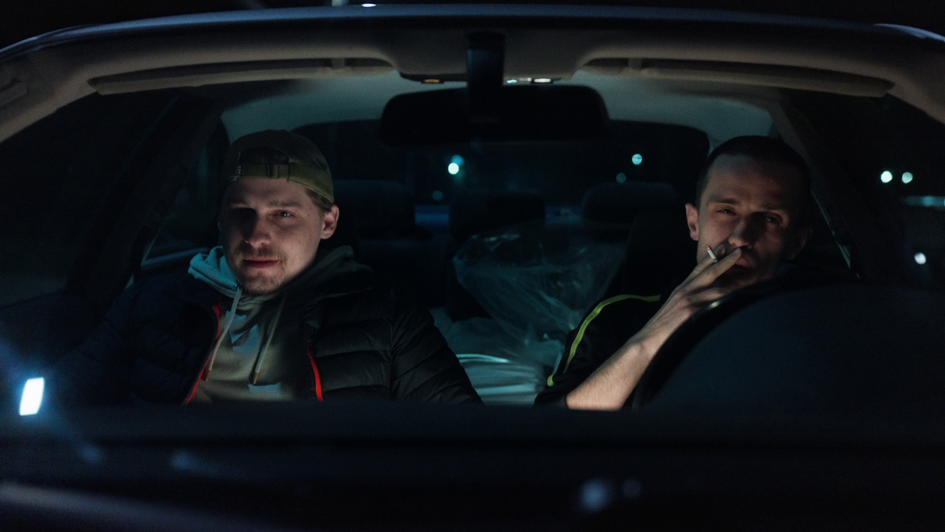 Two men sitting in a car at night, one on the left wearing a cap and jacket, looking serious, and the other on the right smoking a cigarette, with a plastic bag in the back seat.