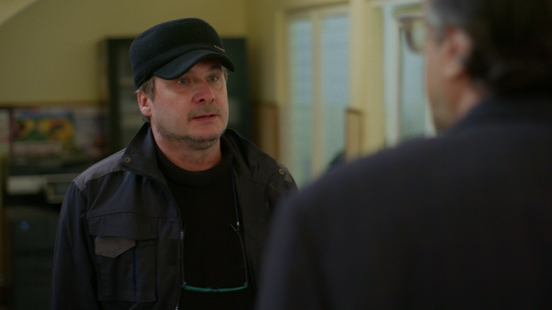 A man wearing a black cap and black jacket standing and talking to another person in an indoor setting.