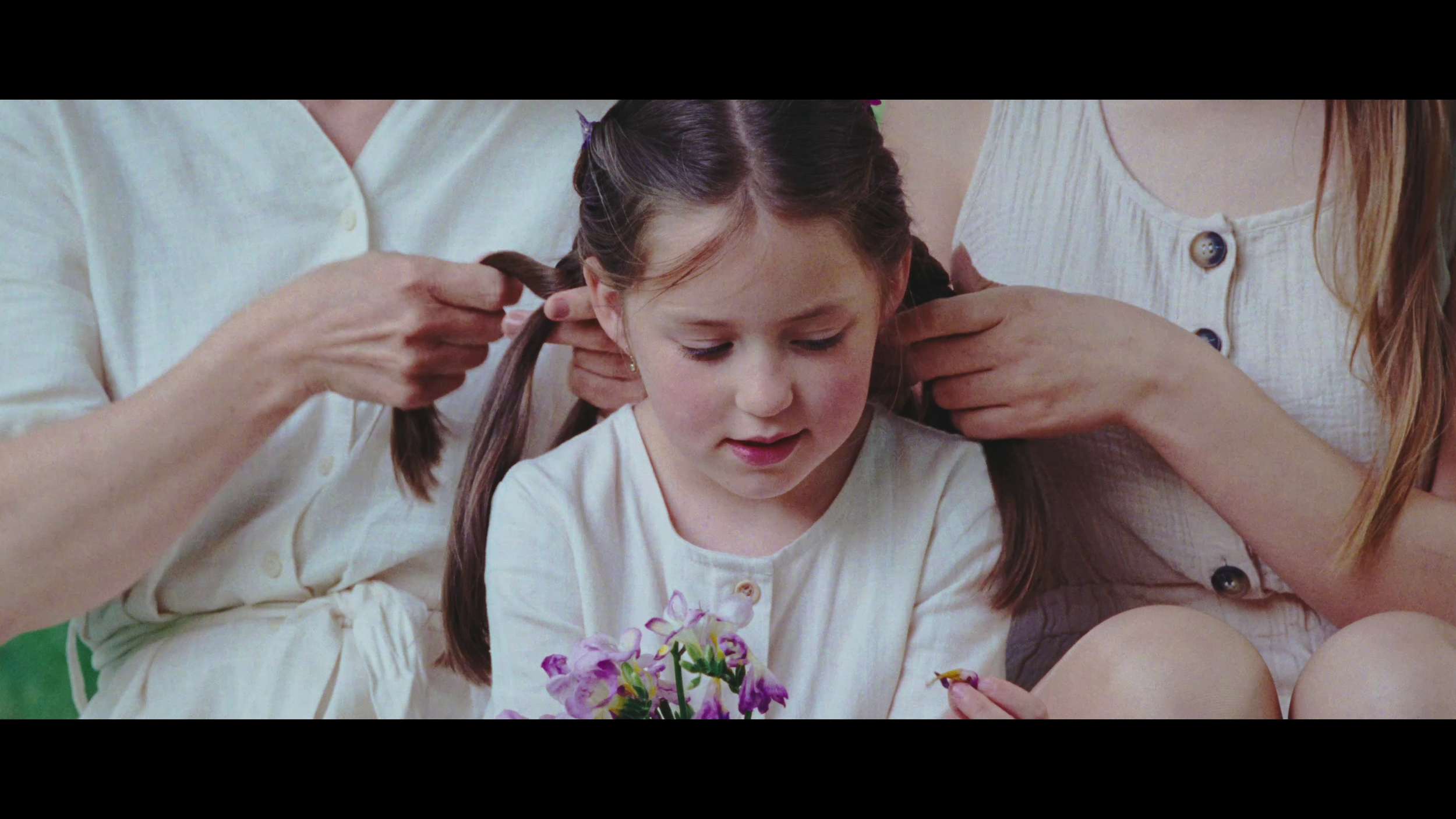 A young girl with long brown hair sitting between two women, getting her hair styled. The girl is holding a small bouquet of purple and white flowers and is looking down at them. The women are helping her with her hair, one holding a section and the other adjusting it.