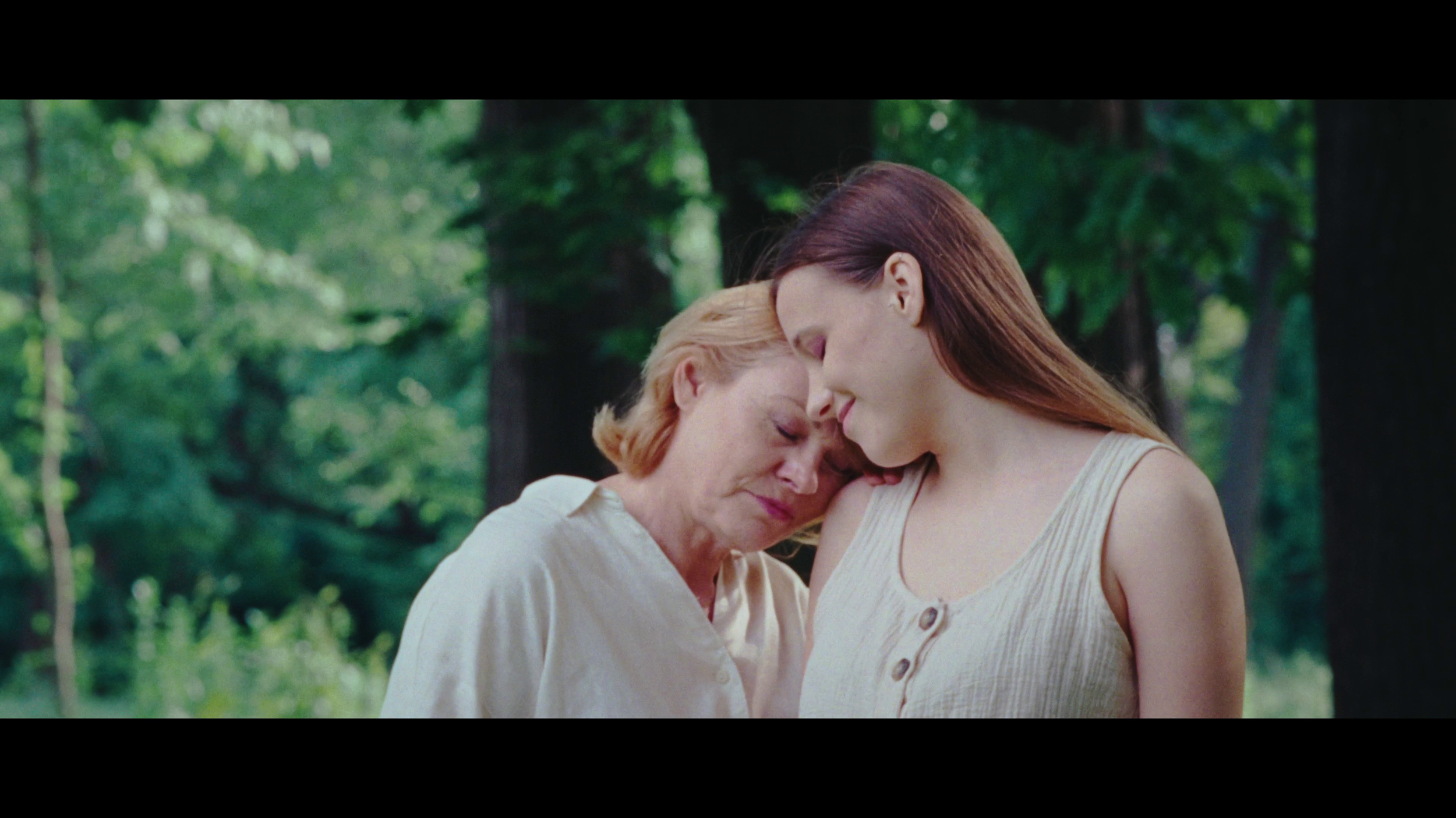 Two women, one older with red hair and one younger with long brown hair, are gently touching foreheads and smiling in a lush, green forest.