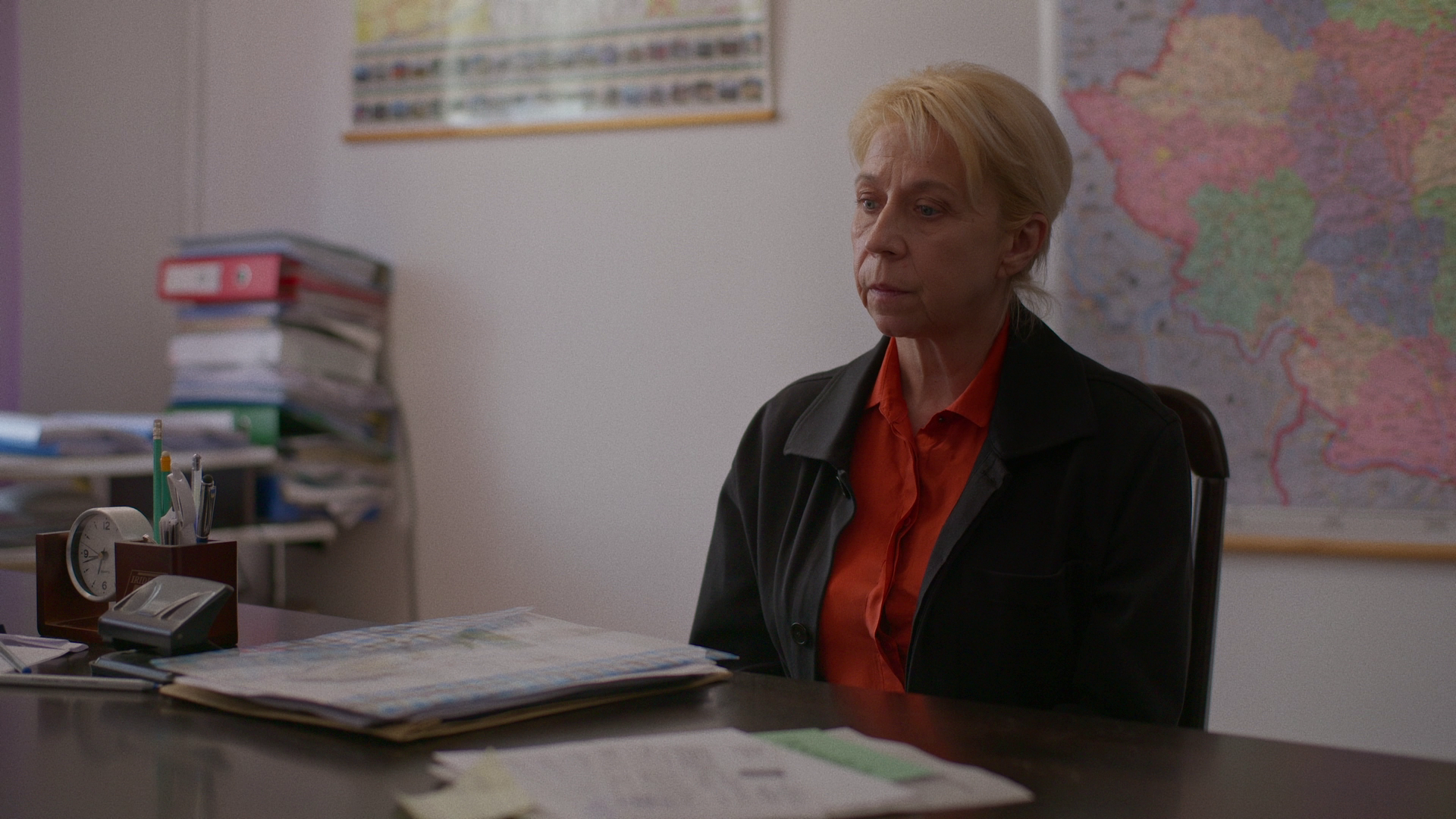 A middle-aged woman with blonde hair sitting at a cluttered desk in an office, with a world map on the wall behind her.