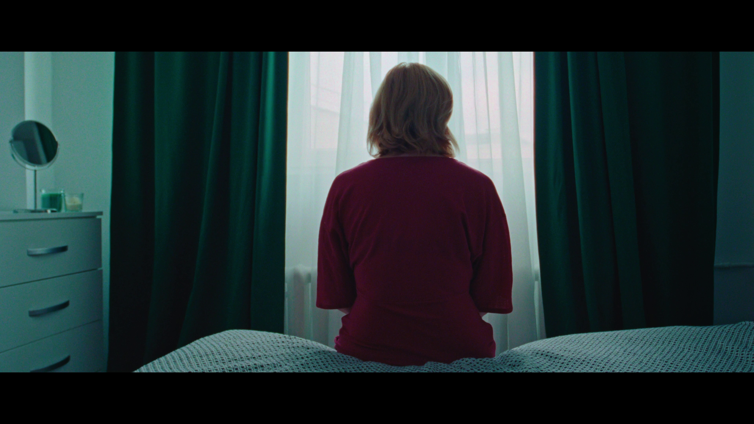 A woman with short brown hair sits on a bed facing a window with white curtains and dark green drapes.
