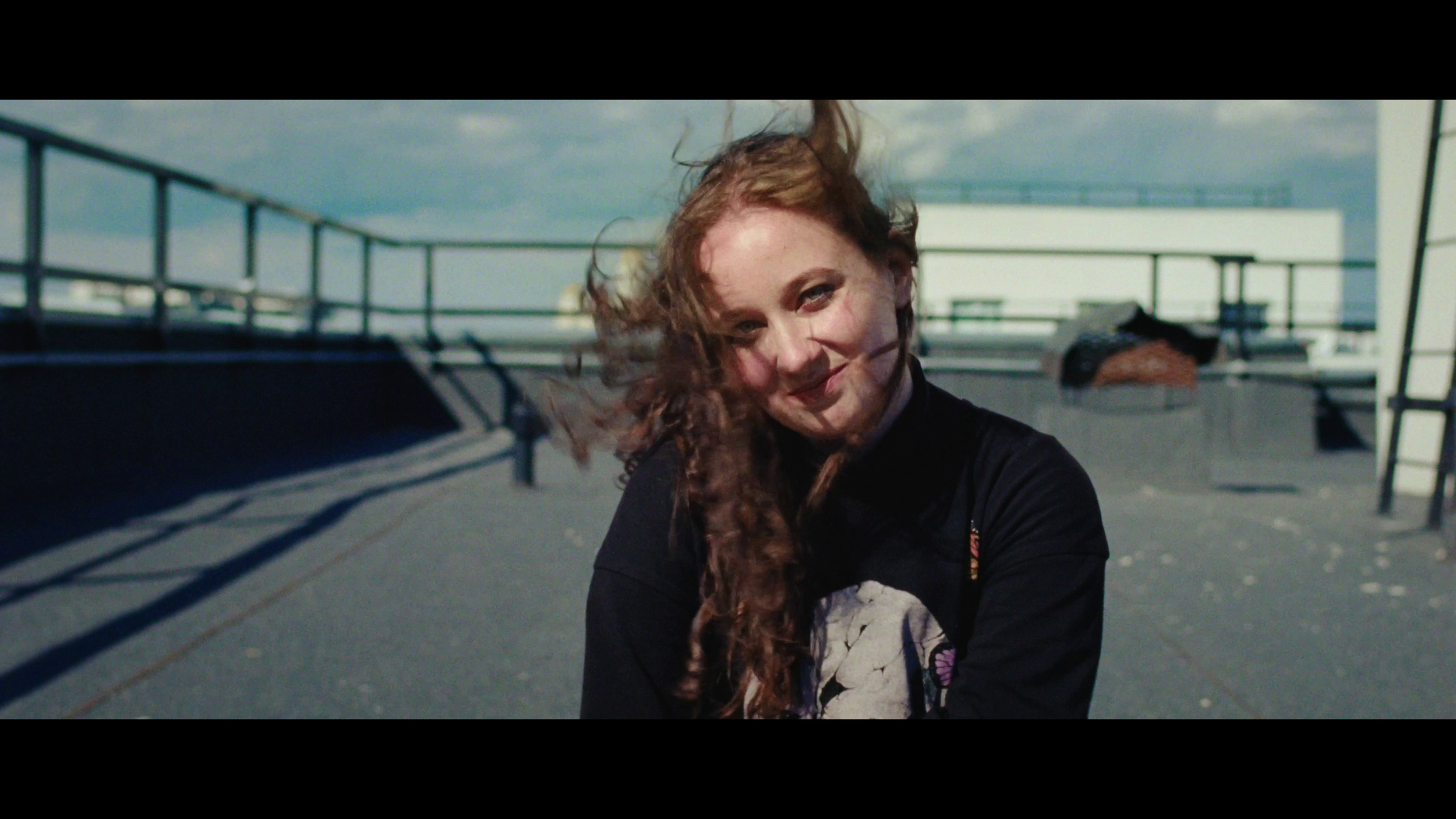 A woman with long curly red hair on a rooftop with a blue sky, smiling and looking into the camera.