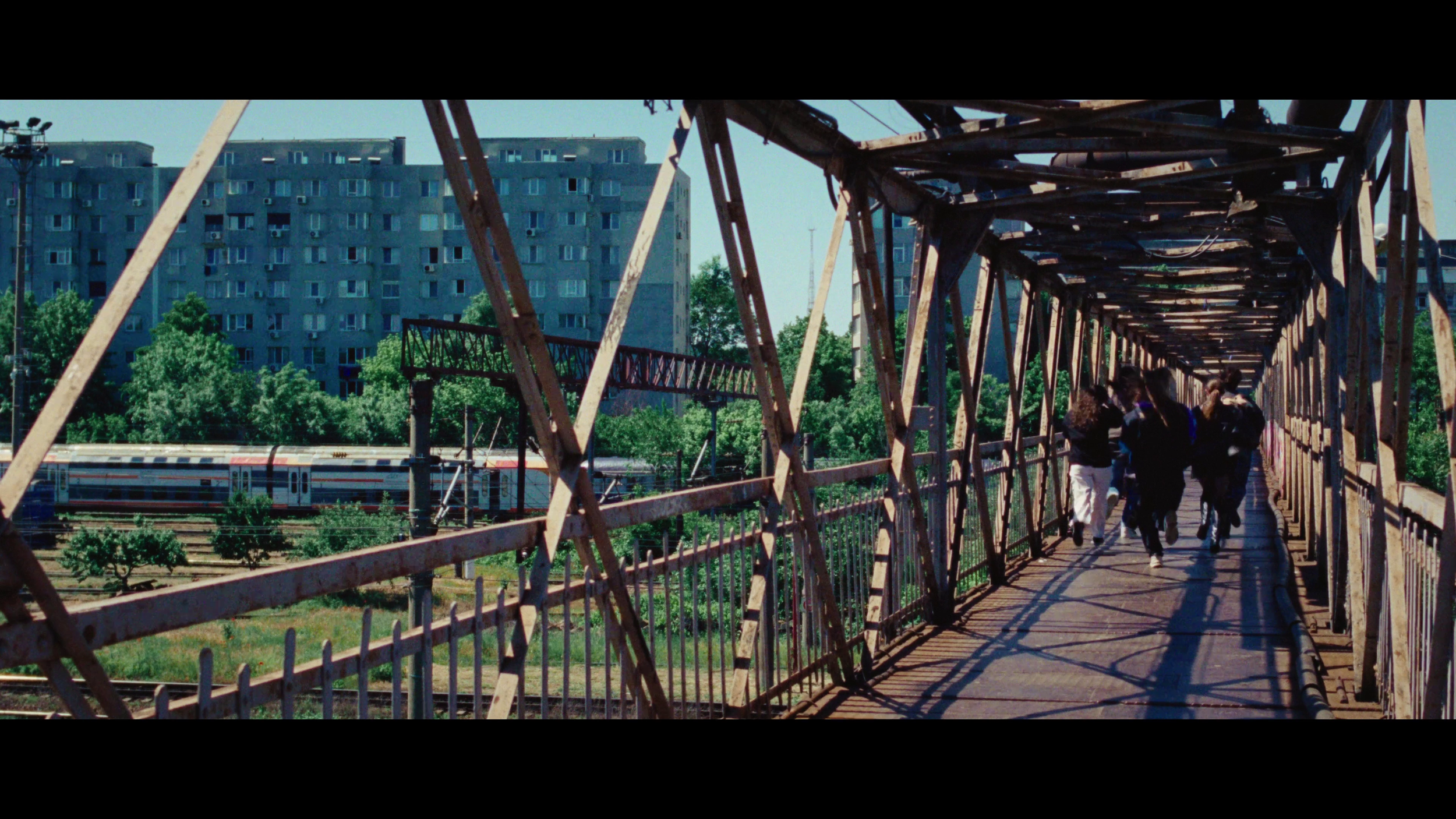 People walking inside a narrow, rusted metal pedestrian bridge over a train tracks with a train passing below, city buildings and green trees in background.