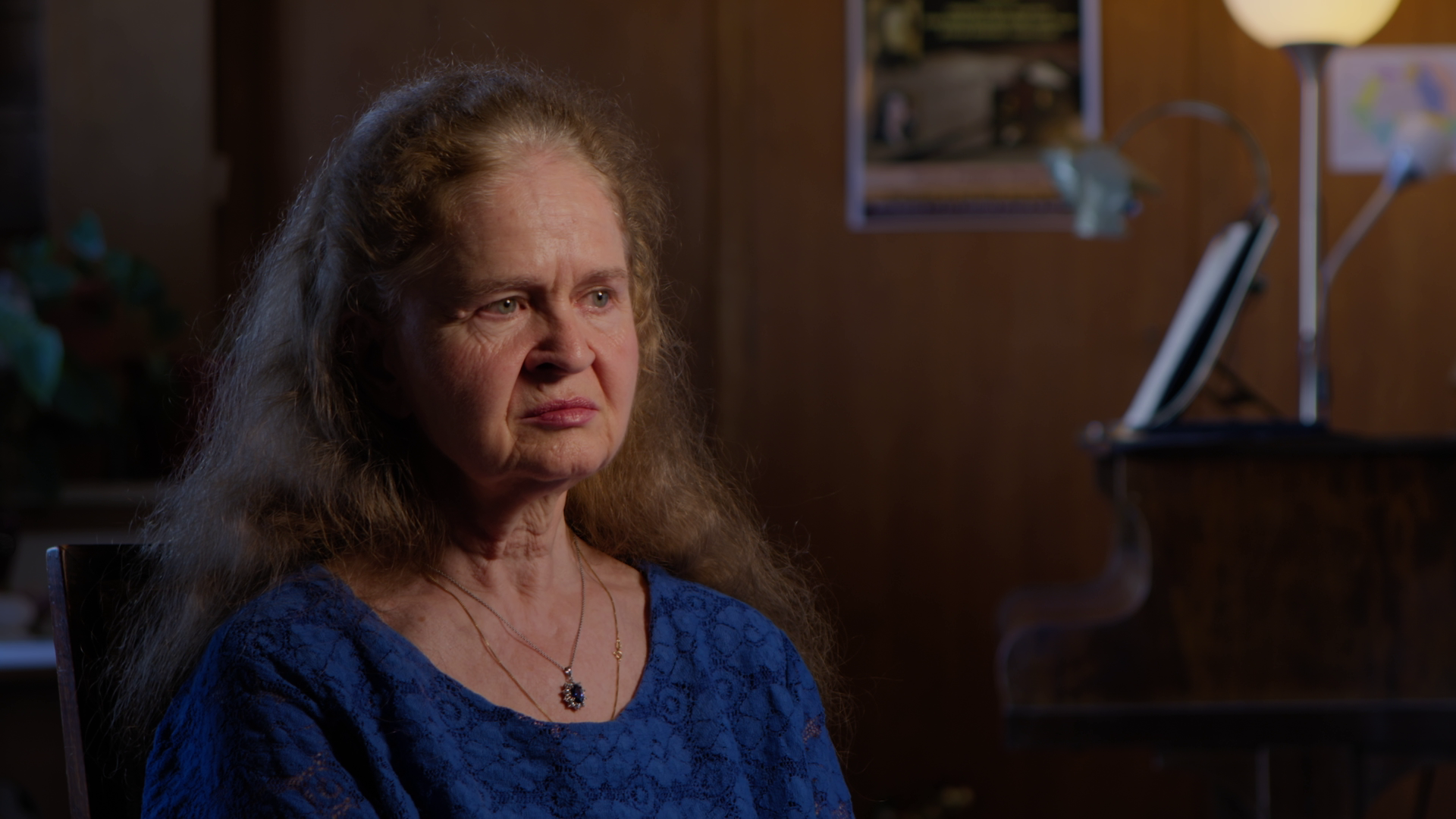 An older woman with long, wavy gray hair and light skin, wearing a blue lace top and a necklace, sitting indoors with a serious expression. In the background, there is a wooden wall, a poster, a flower-shaped lamp, and a piano.