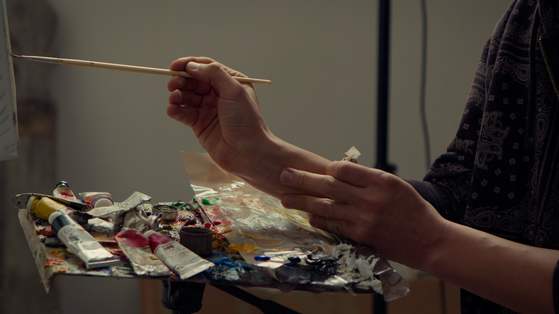 Artist using a brush to paint on a hand model's arm, with a cluttered palette of paints and brushes on a table.