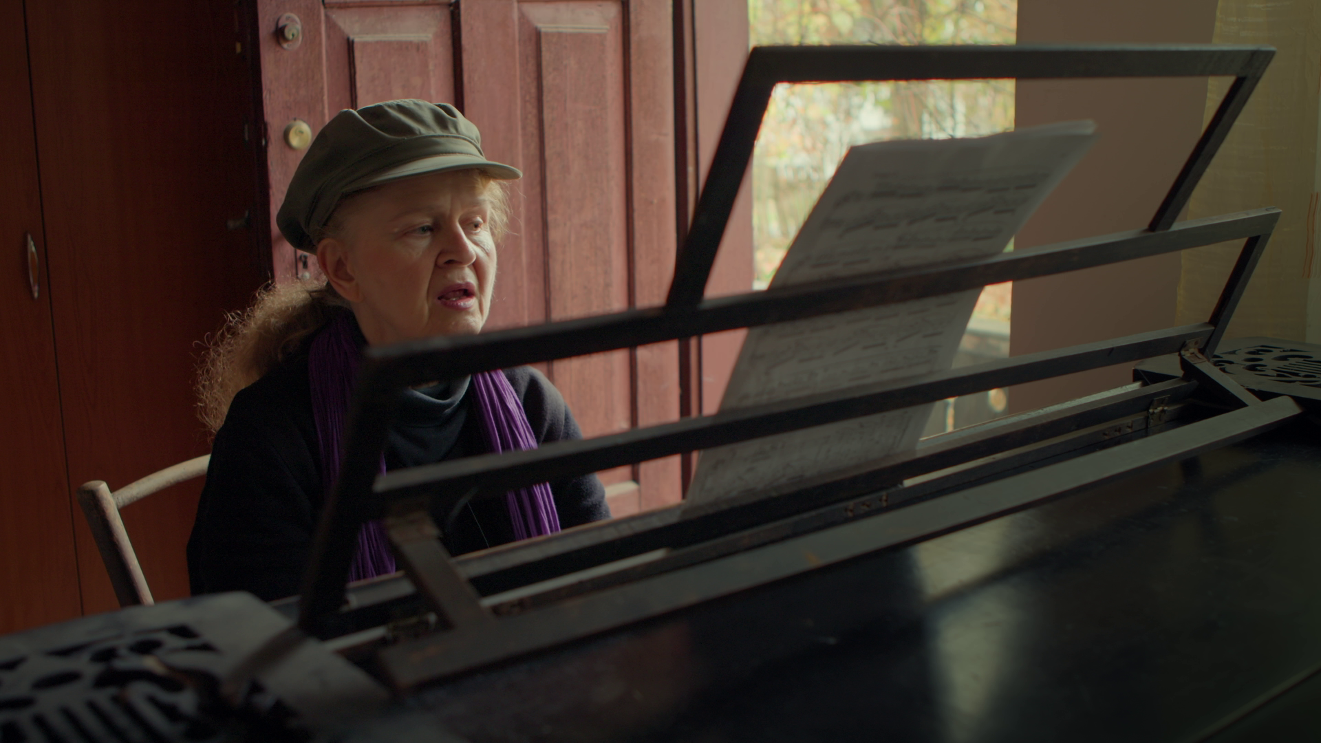 Older woman with long hair wearing a beige hat and purple scarf sitting at a piano in a room with wooden door and window.