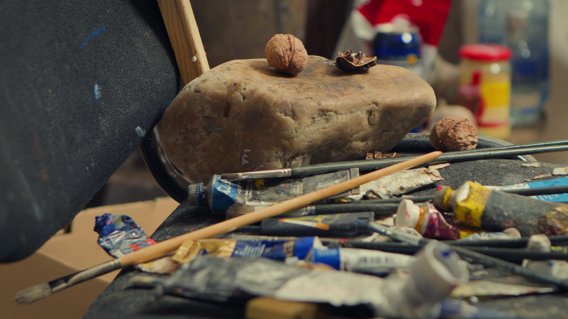 A cluttered artist's workspace with a large stone, walnuts, paint tubes, brushes, and art supplies scattered on a table.