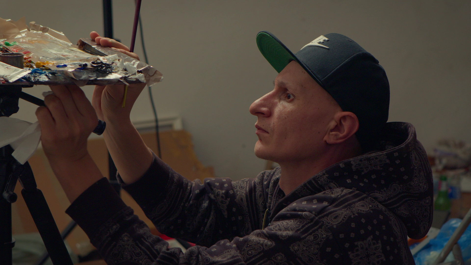 A man wearing a black cap and a patterned hoodie concentrating while painting on a palette of various colors.