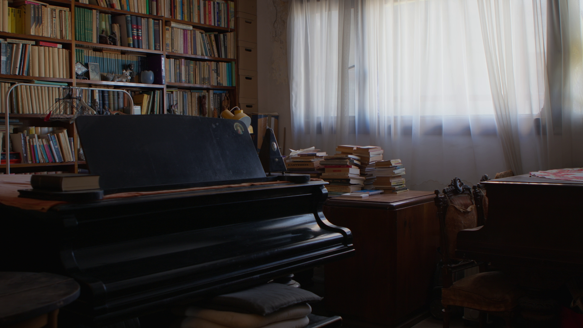A dimly lit room with a black piano covered in books and papers, wooden furniture, a large bookshelf filled with books, and a window with sheer curtains allowing natural light to filter in.