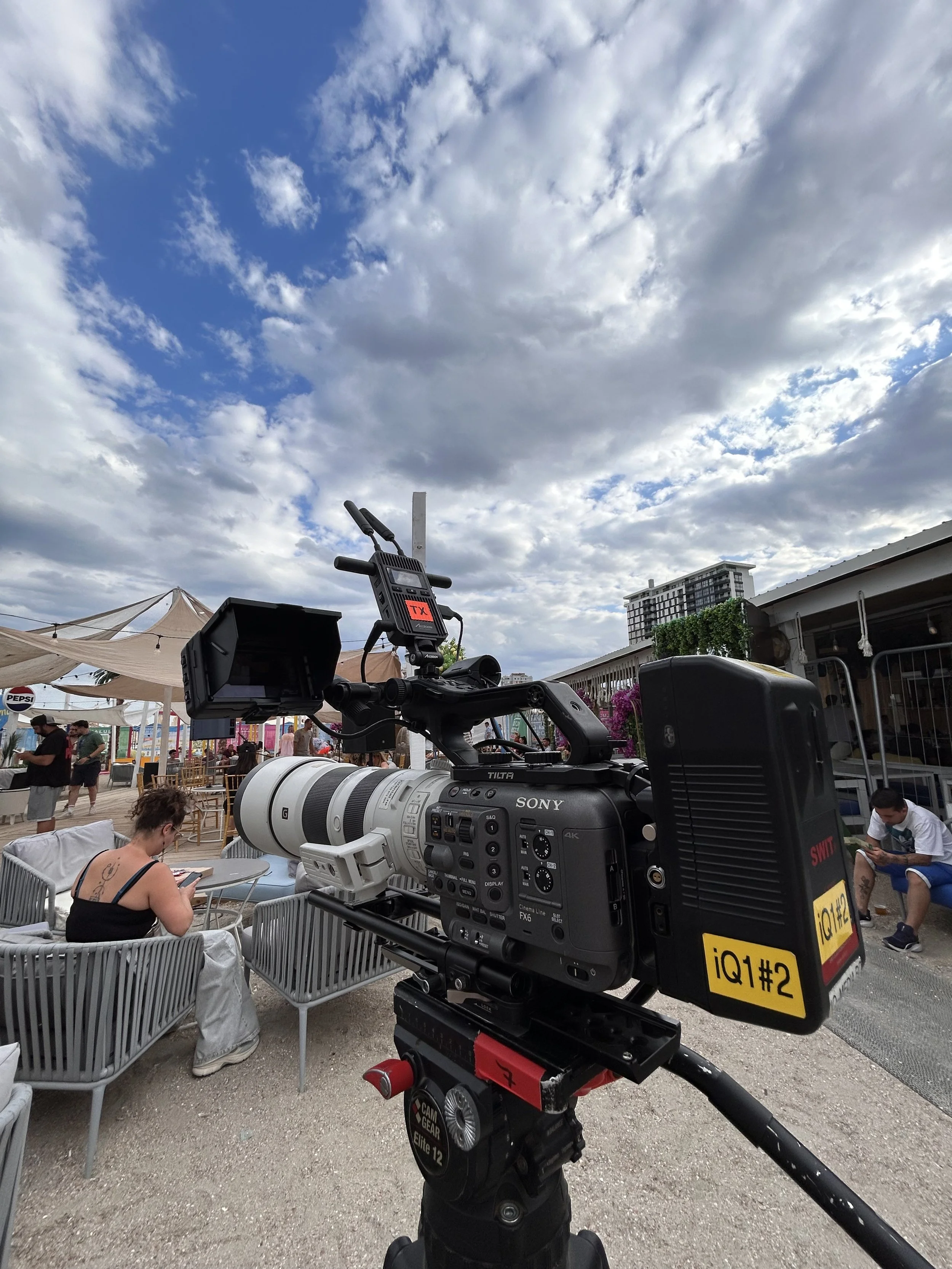 A professional video camera on a tripod at an outdoor event with people in the background, some sitting and others walking, under a partly cloudy sky.