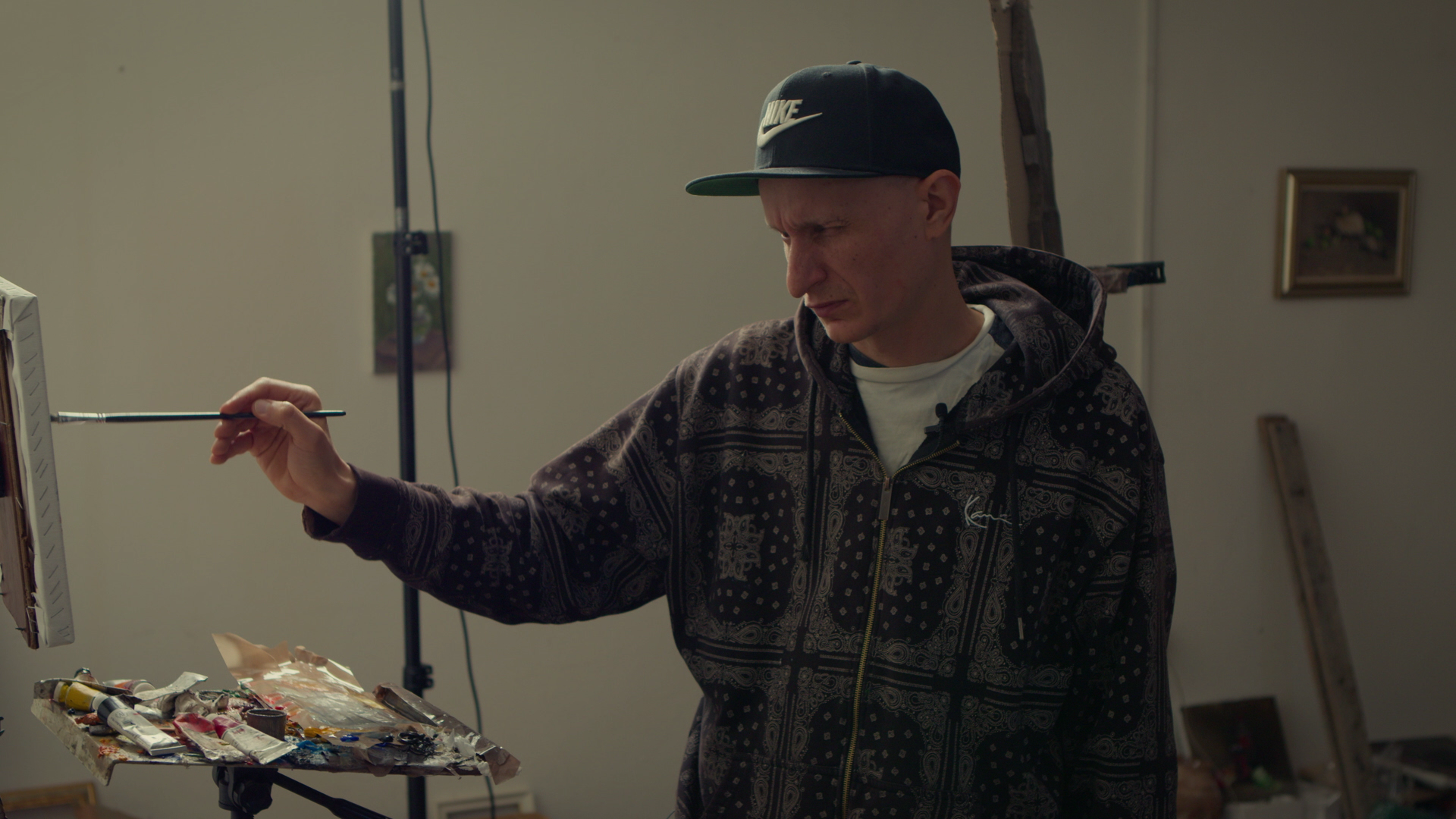 A man wearing a black Nike cap and a patterned hoodie is painting on a canvas setup in an indoor studio, with a palette of paint and tubes on a table in front of him.
