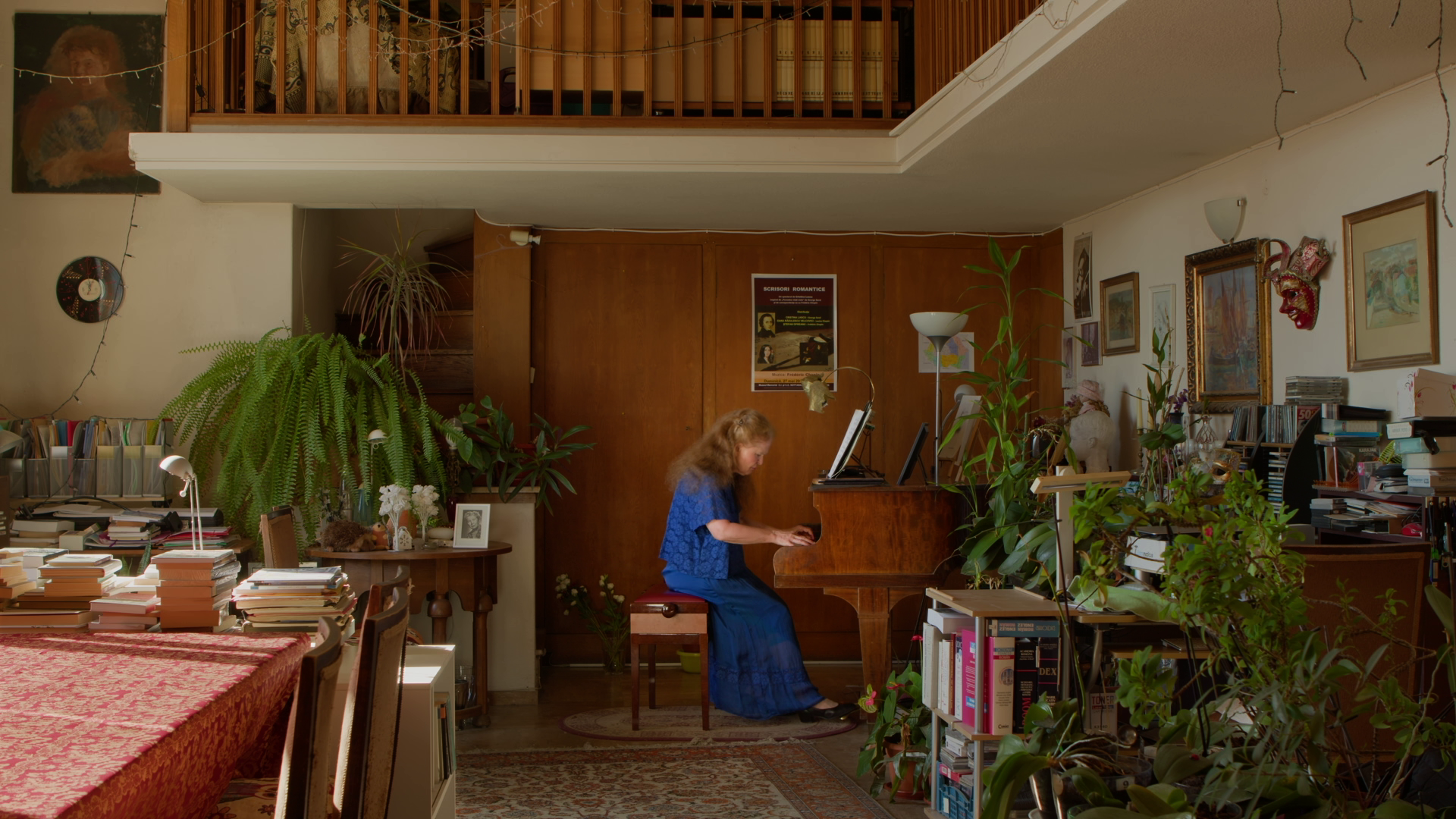A woman with curly red hair in a blue dress playing the piano in a warmly decorated living room filled with plants, books, artwork, and decorative objects.