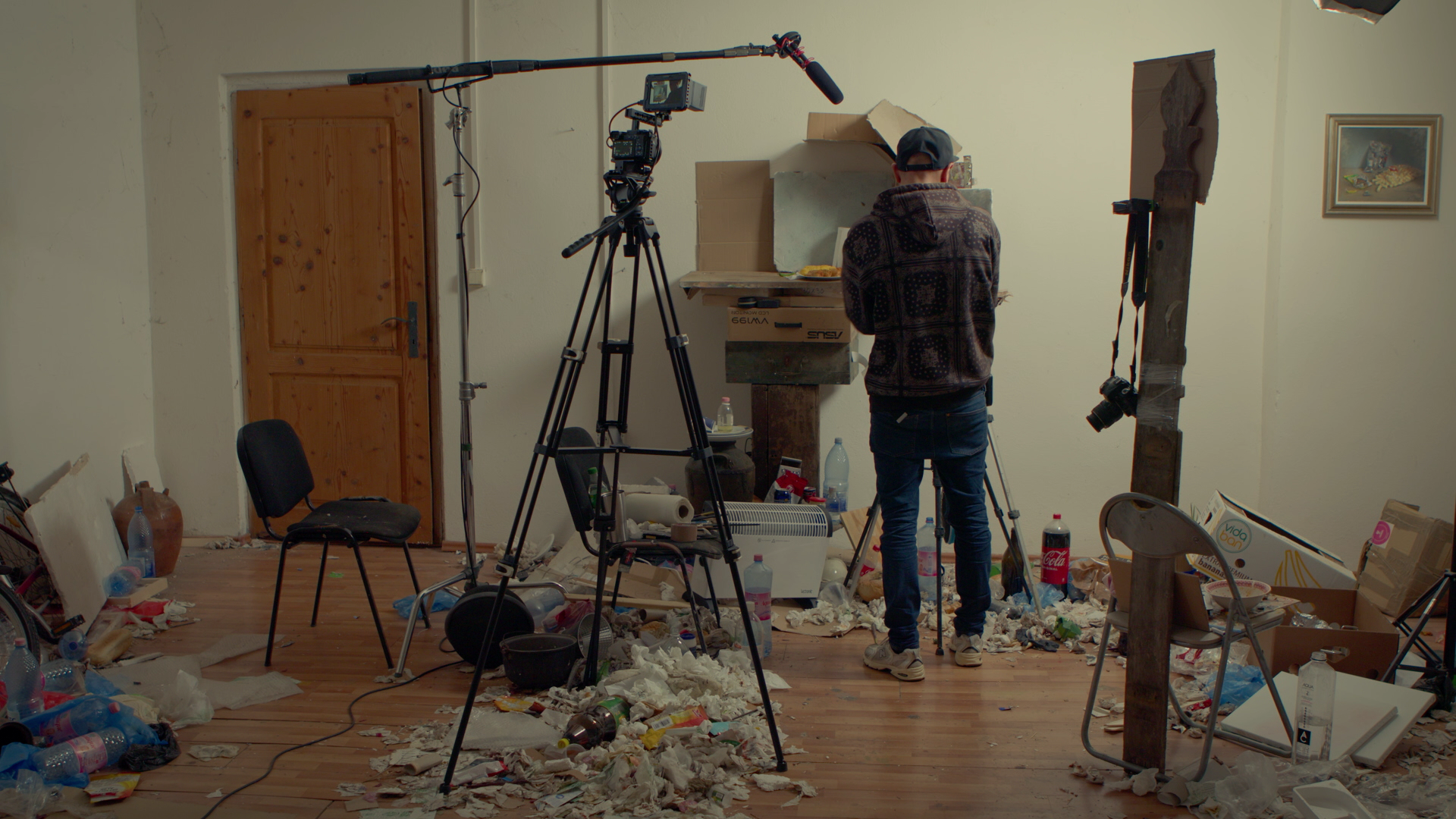 A person filming in a cluttered, messy room with debris, chairs, camera equipment, and trash scattered across the floor.