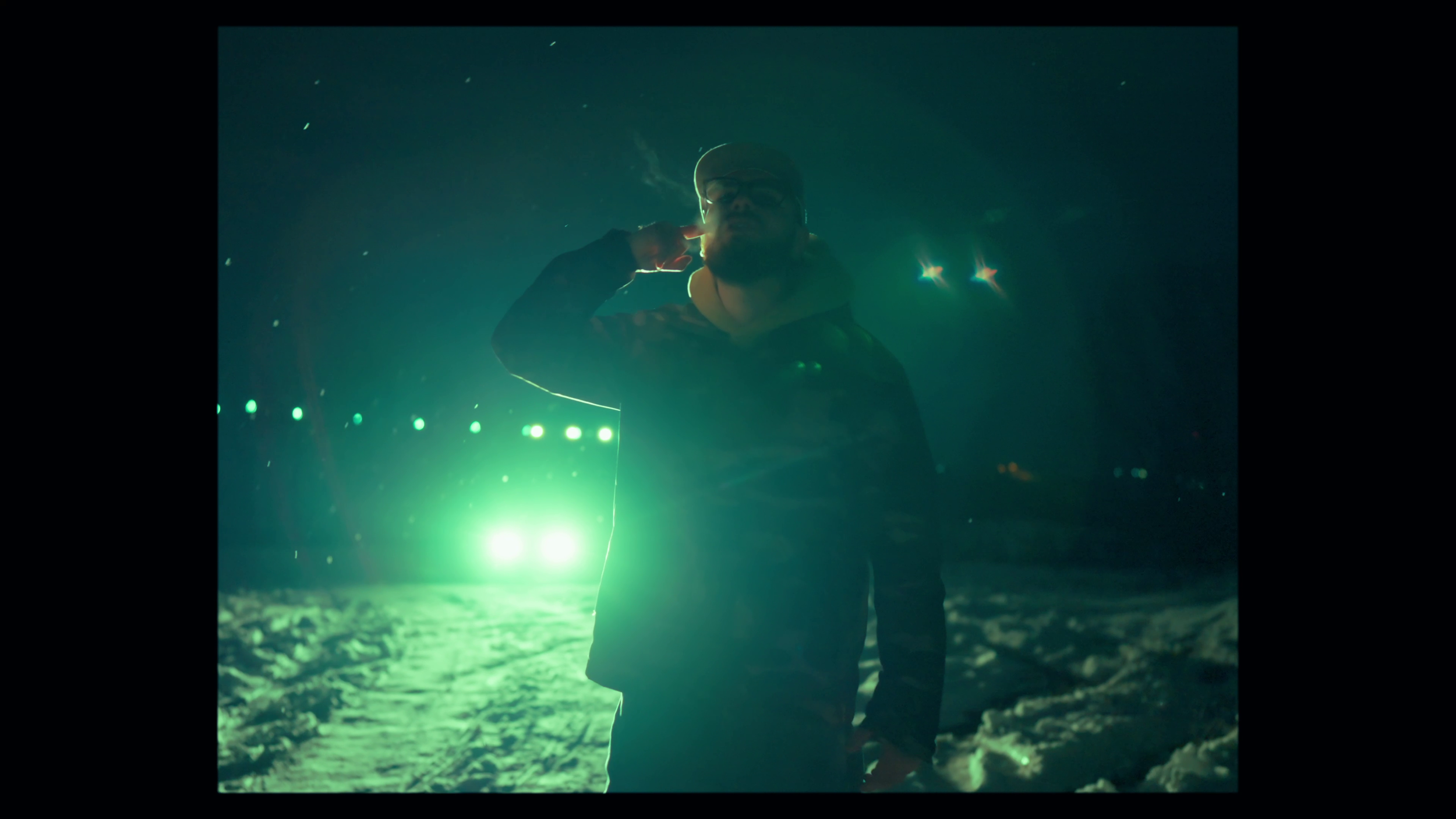 A man standing outdoors at night in a snowy landscape, illuminated by bright green and white lights in the background, wearing a dark jacket and cap, with his hand near his ear.