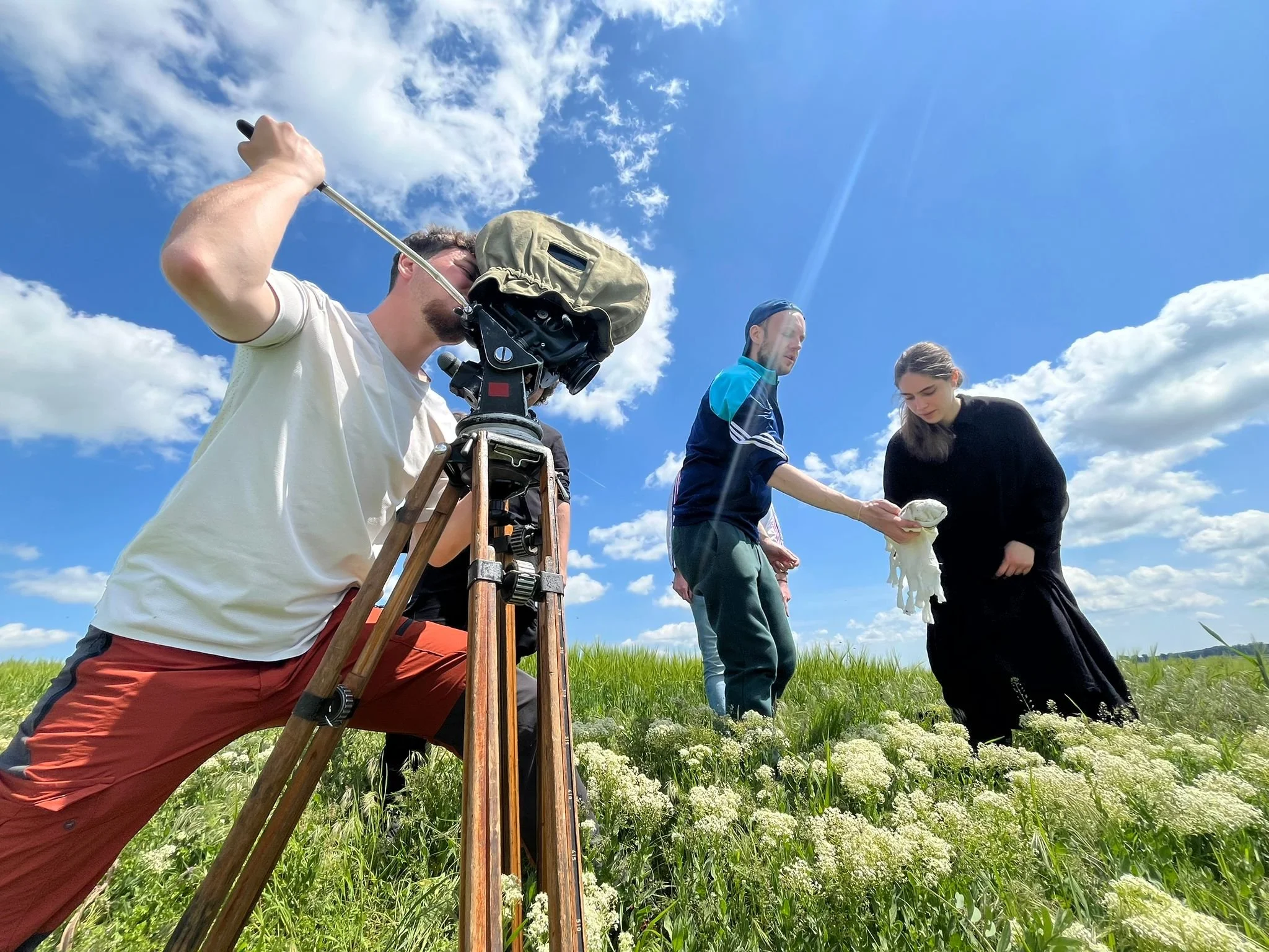 A film crew shoots a scene outdoors in a grassy field with white flowers, under a blue sky with clouds. A man operates a camera on a tripod while two women, one holding a cloth or prop, interact with each other.