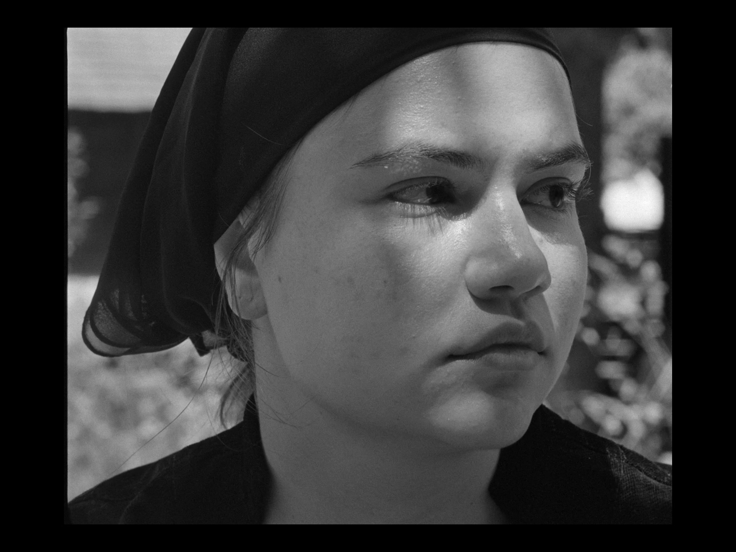 Close-up black and white portrait of a young woman's face with a scarf on her head, outdoors with blurred trees in the background.
