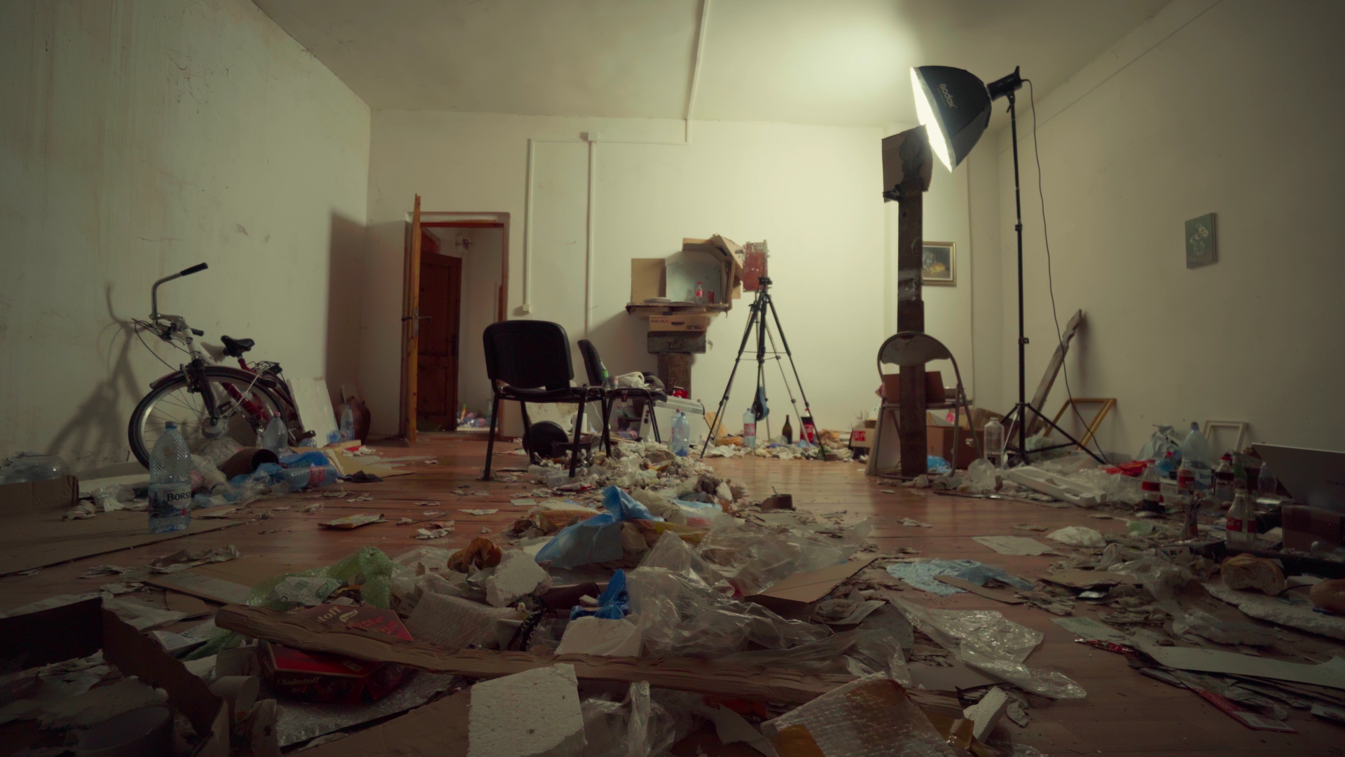 Disorganized room with scattered trash, a bicycle, chairs, a tripod, and a lighting setup.