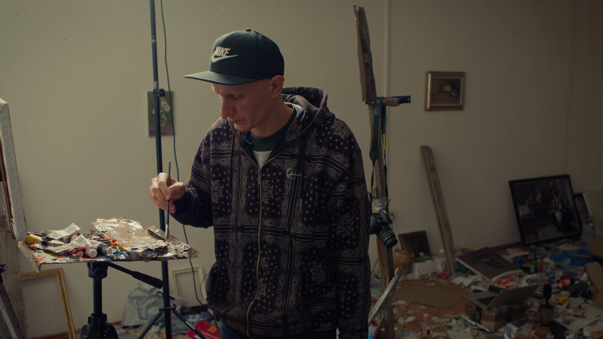 A man wearing a black Nike cap and a patterned hoodie standing at an art easel with a palette of paint, painting on a canvas in a cluttered room with scattered items and photos.