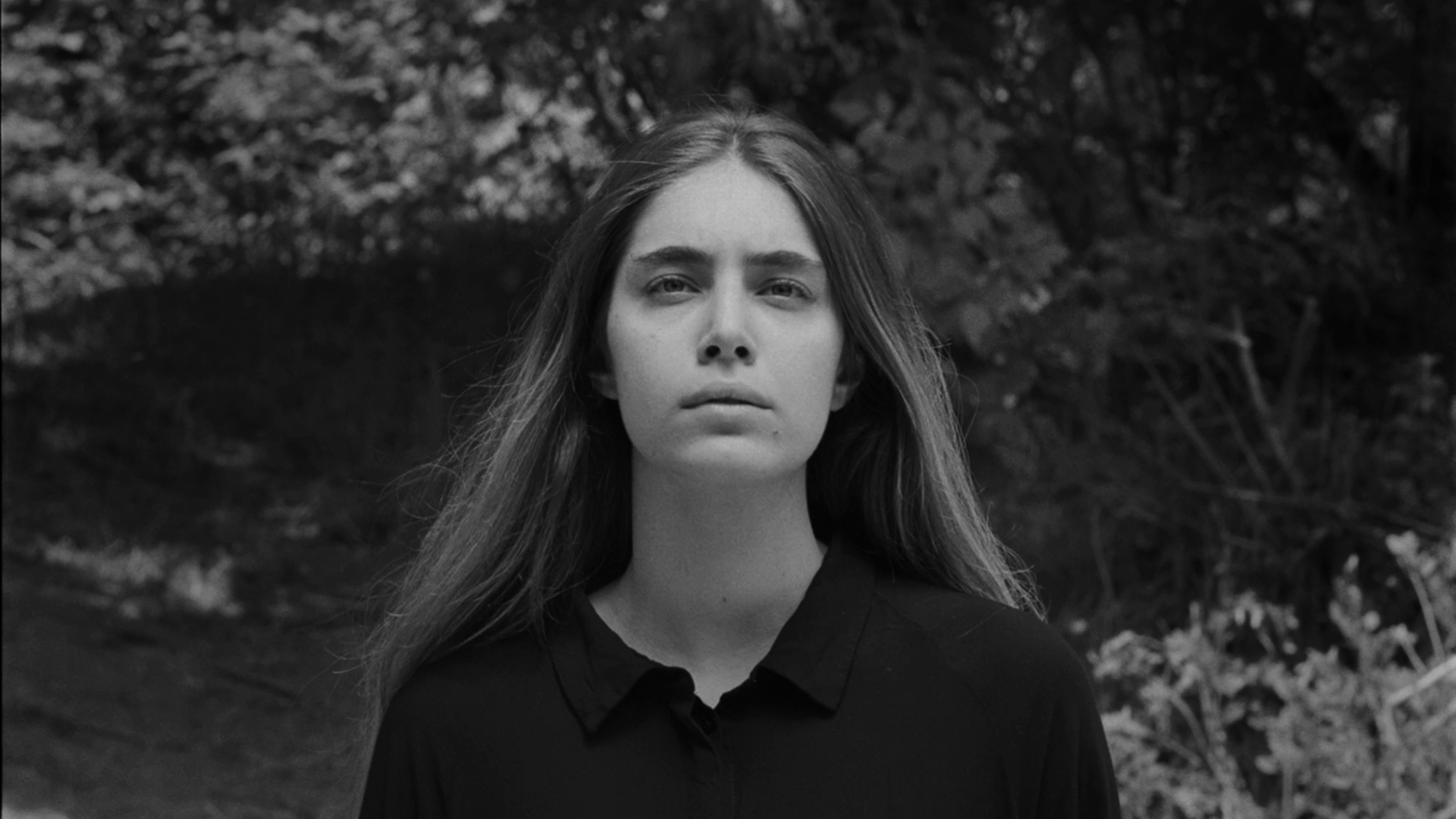 Black-and-white photo of a woman with long hair, wearing a dark shirt, standing outdoors with trees and foliage in the background.