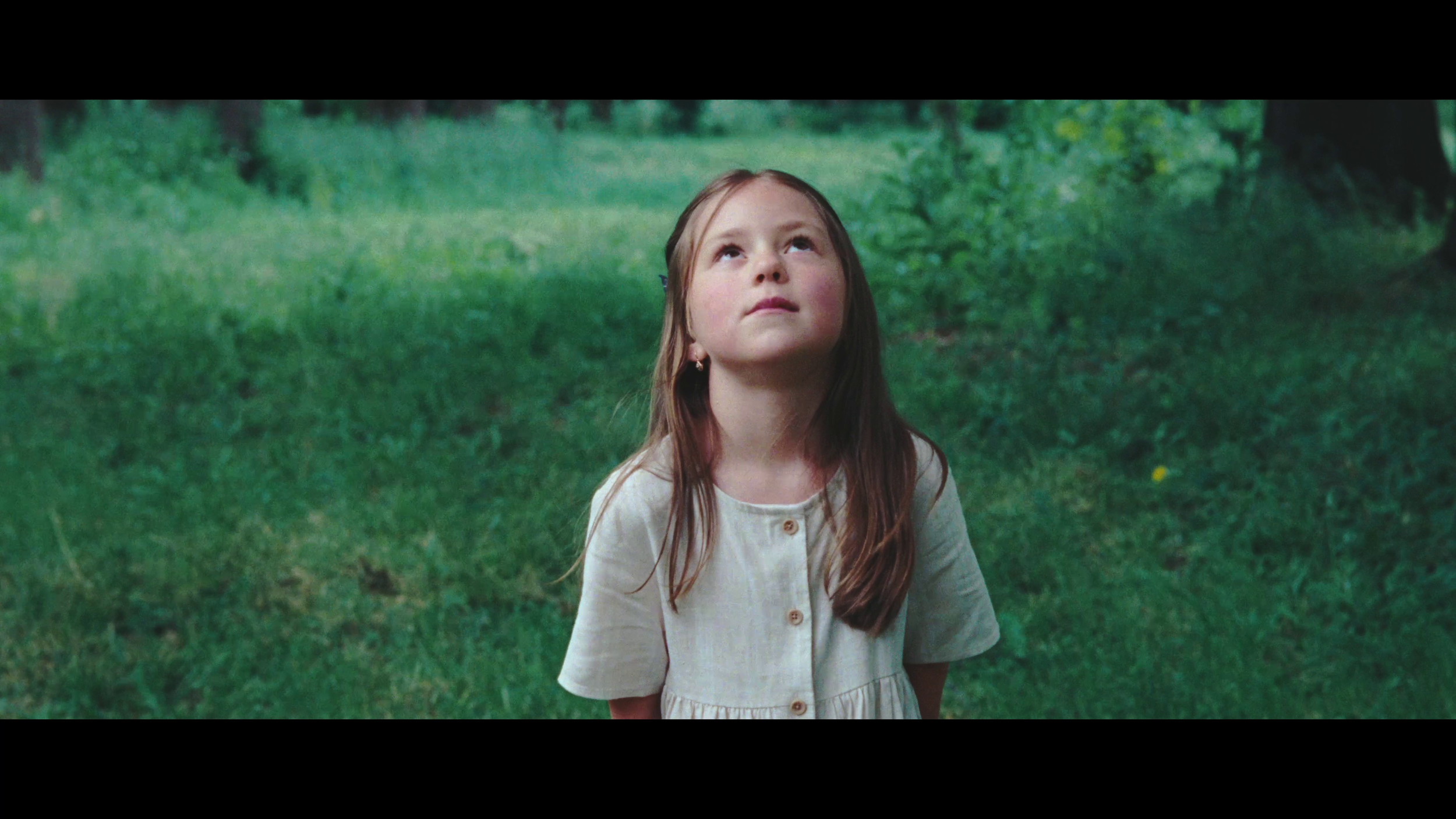 A young girl with long brown hair, wearing a beige dress, looking upward in a green outdoor setting with grass and trees.