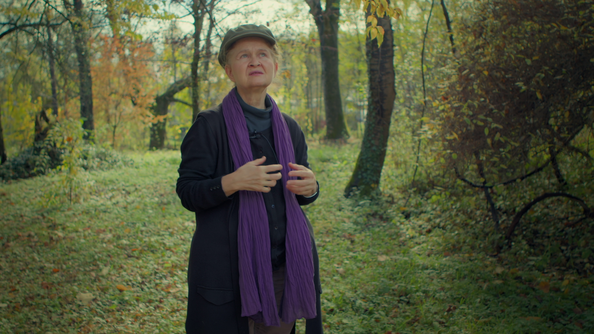 An elderly woman with a purple scarf and a gray hat standing in a wooded forest during fall, looking upward with a thoughtful expression.