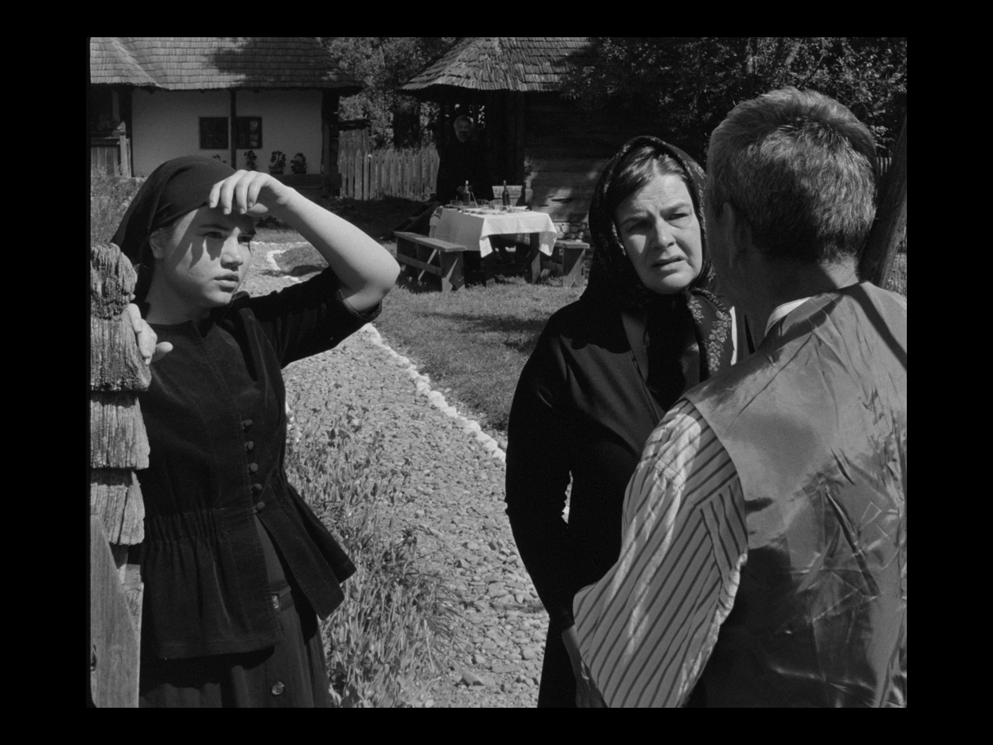 Black and white photo of three people having a conversation outdoors in a rural setting with traditional wooden houses. A girl wearing a headscarf shields her eyes from the sun while a woman in a headscarf listens to a man with a striped shirt and vest.