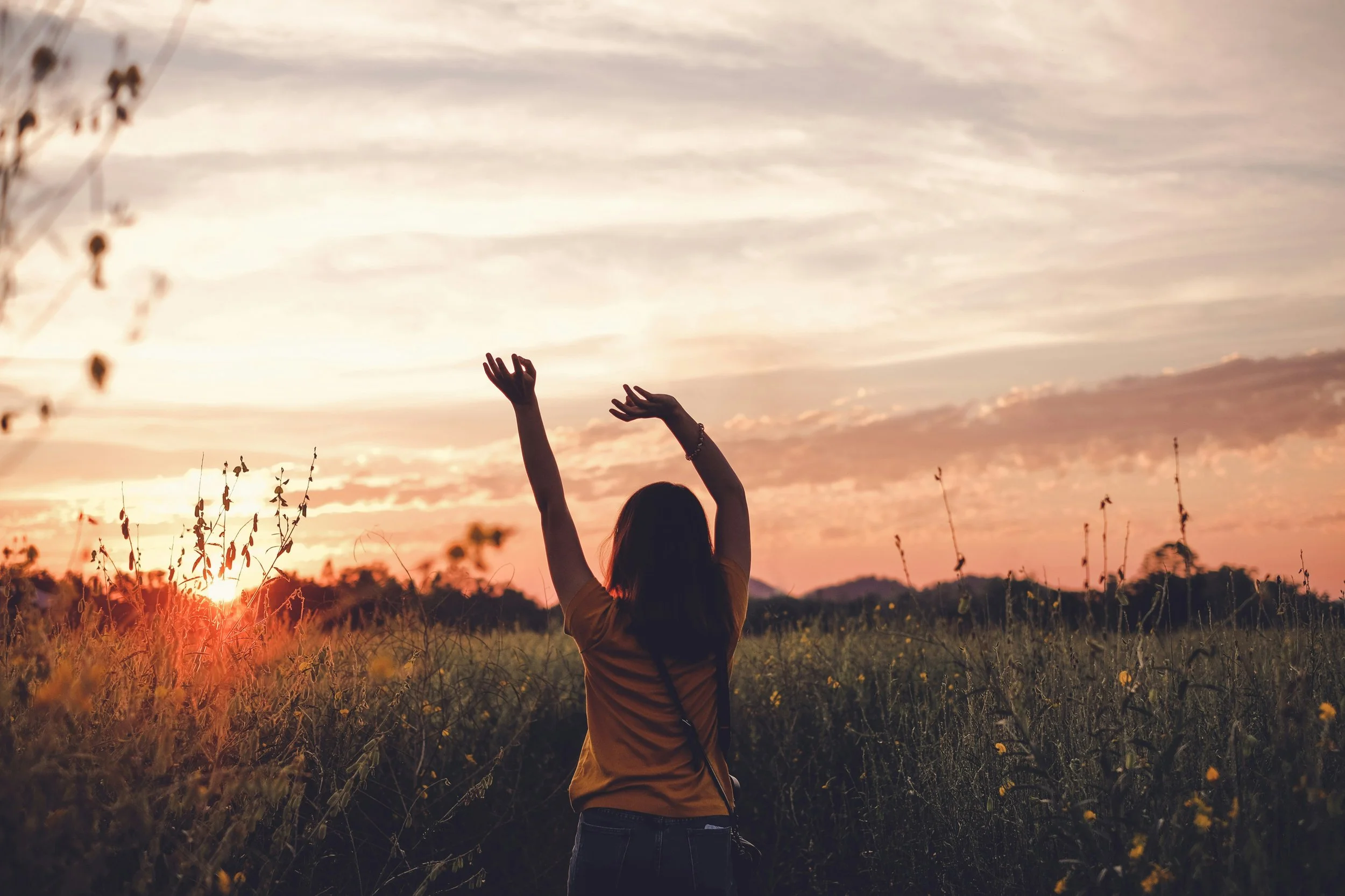 A woman standing in a field at sunset with her arms raised.