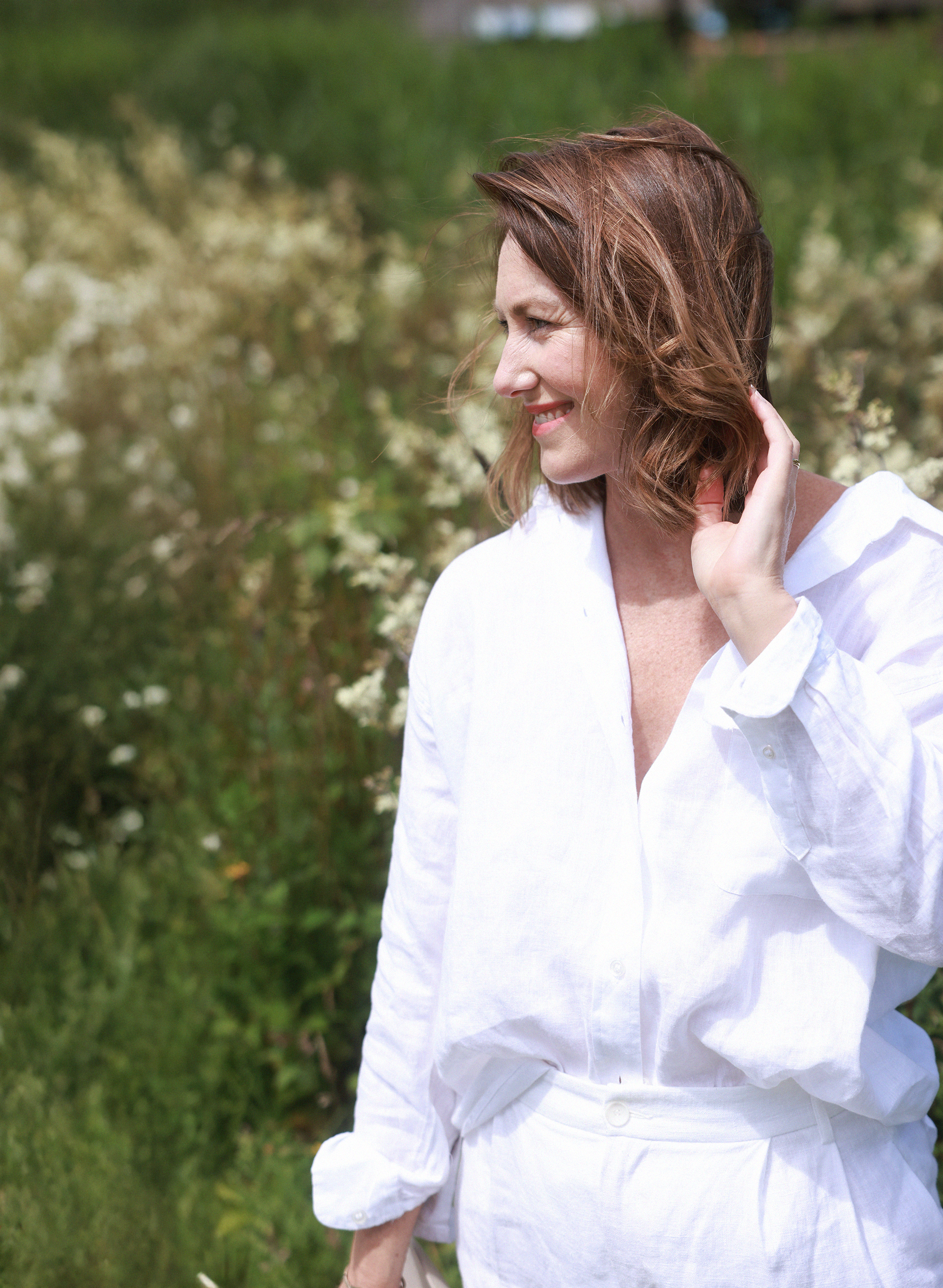 A woman with shoulder-length auburn hair smiling and touching her neck, standing outdoors in a natural setting with greenery and white flowering bushes in the background.