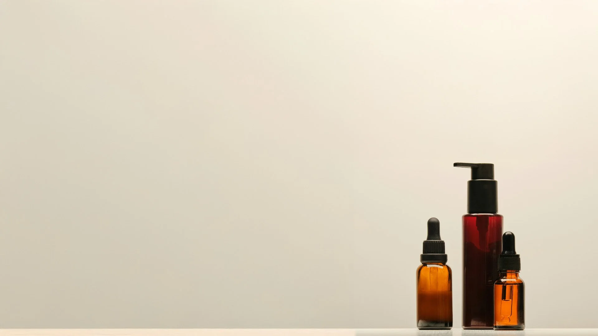 Three skincare bottles with dropper and pump tops are arranged on a surface against a plain, light background.