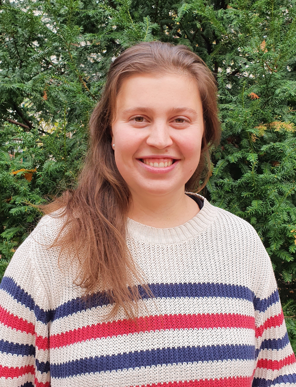 Junge Frau mit braunen Haaren, die einen Strickpullover mit roten, weißen und blauen Streifen trägt, vor einem grünen Busch.