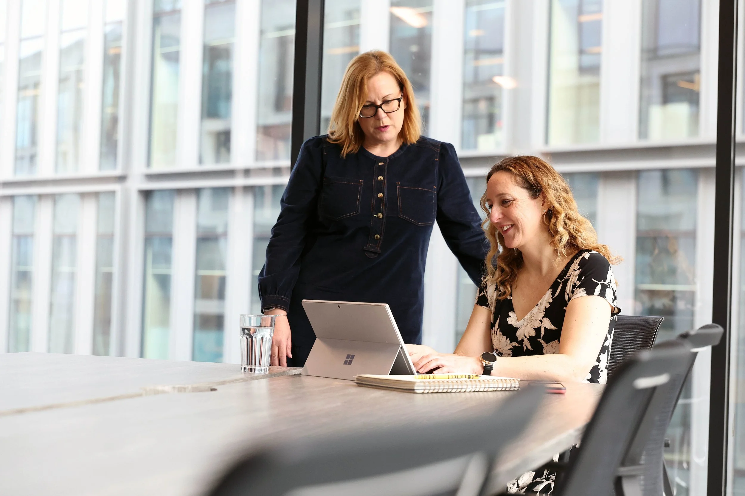 Two women in a modern conference room, one standing and the other sitting at a table, looking at a laptop together, with large windows in the background.