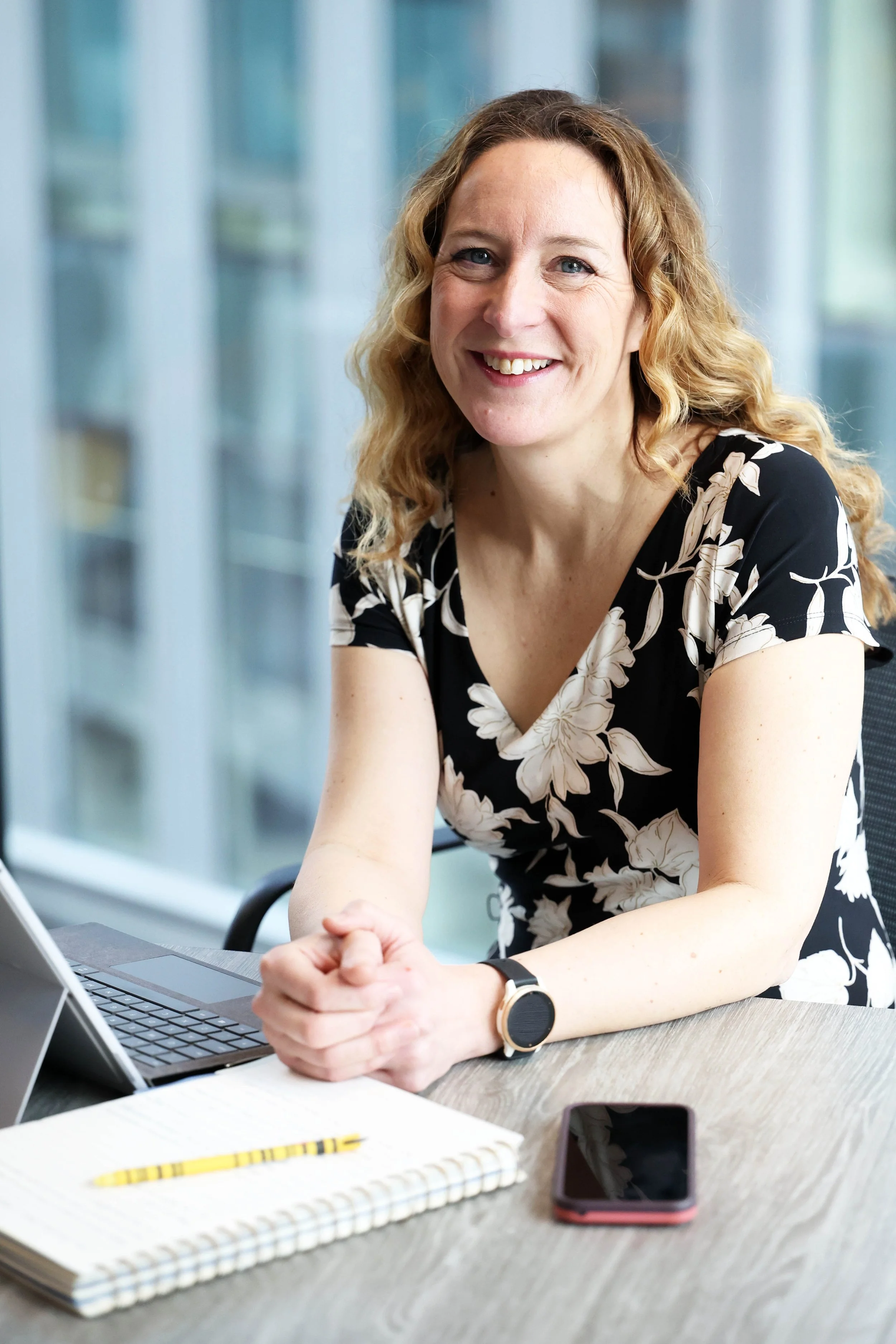 A woman with curly blonde hair smiling while sitting at a desk with a laptop, notebook, pen, and smartphone in a modern office with large windows.