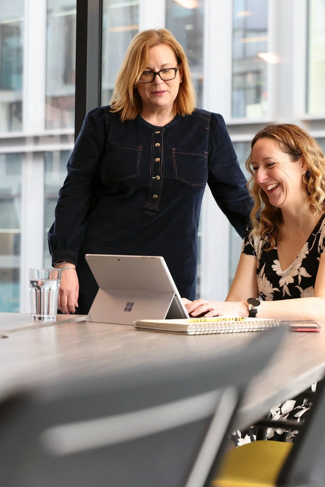 Two women are in a modern office, one standing and the other sitting, working on a laptop together, with a glass of water and a notebook on the table.