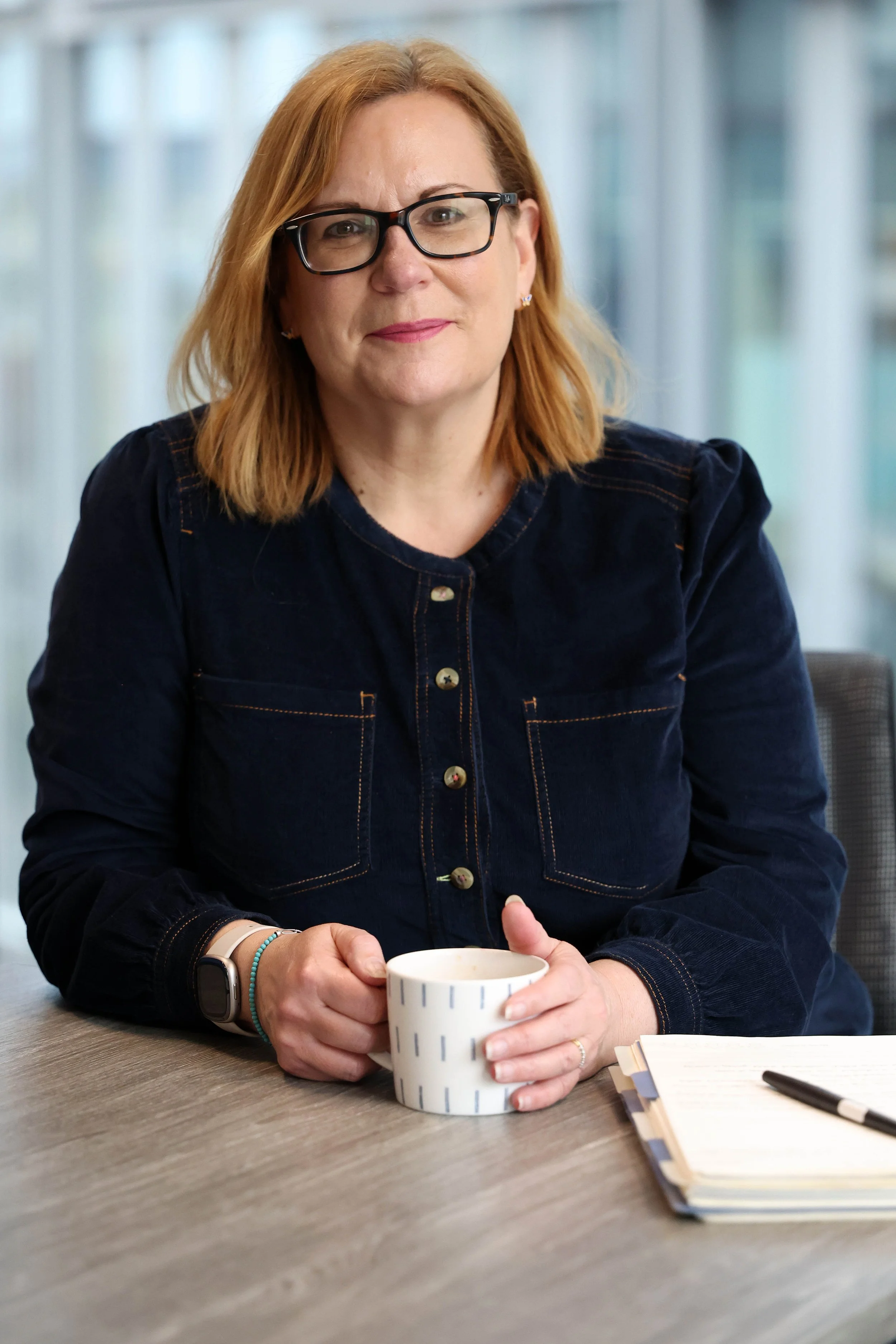 A woman with shoulder-length red hair and glasses sitting at a table, holding a cup of coffee, with a notebook and pen in front of her.