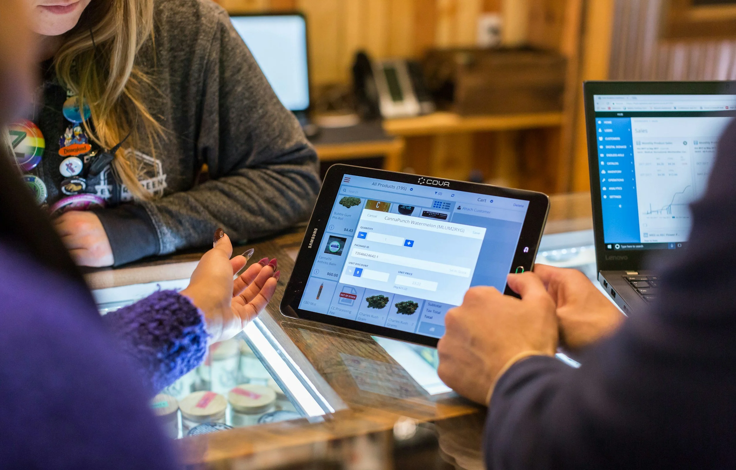 People discussing and using a tablet and laptop at a wooden counter in an indoor setting, with a display case of small items on the counter.